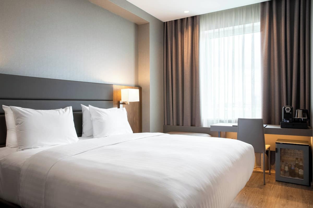 A hotel room with a large bed, a desk with a chair, a coffee machine, and a minibar.