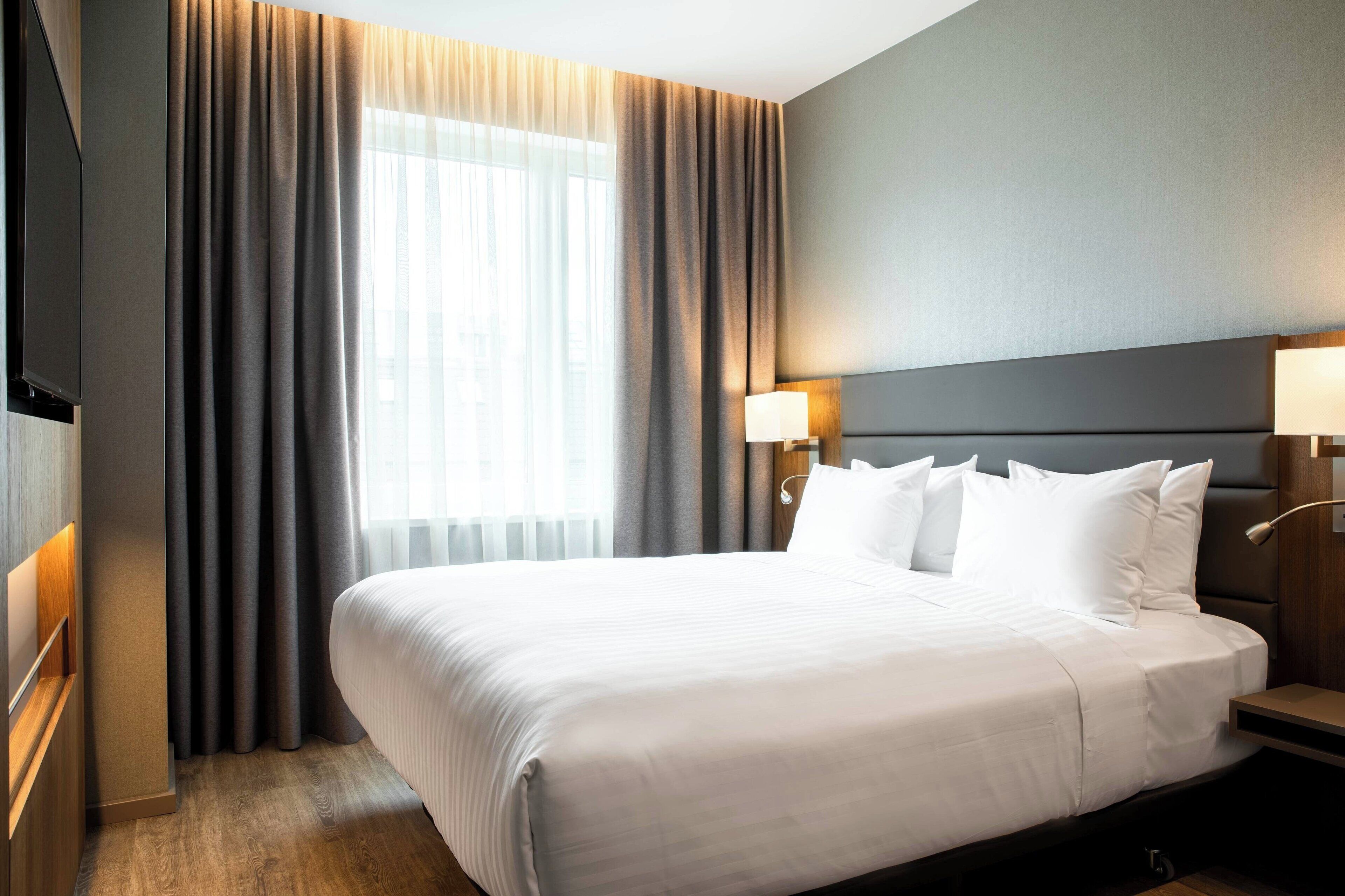 A hotel room with a large bed, a flat-screen TV, a nightstand, and a window with curtains.