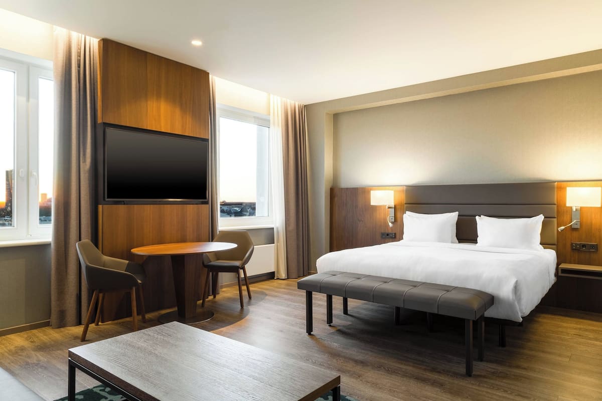 A hotel room with a bed, bedside tables, a TV, a desk with chairs, and a window with curtains.