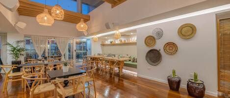 A spacious dining area with wooden tables and chairs, a high ceiling with wooden beams, and pendant lighting.
