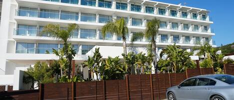 A multi-story hotel with balconies, a parking area, and a fence with greenery.