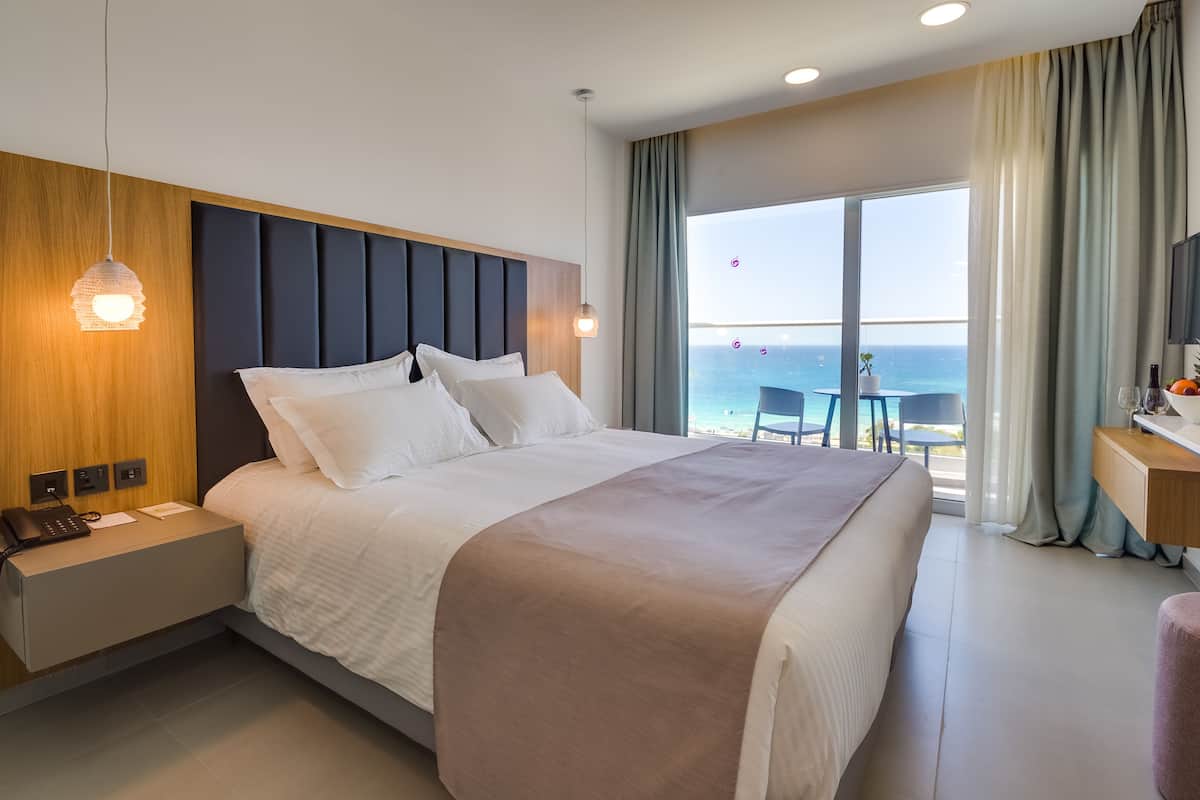 Une chambre d’hôtel moderne avec un grand lit, une table de chevet avec un téléphone, un bureau avec un panier de fruits et des verres, et une vue sur l’océan grâce à de grandes fenêtres.