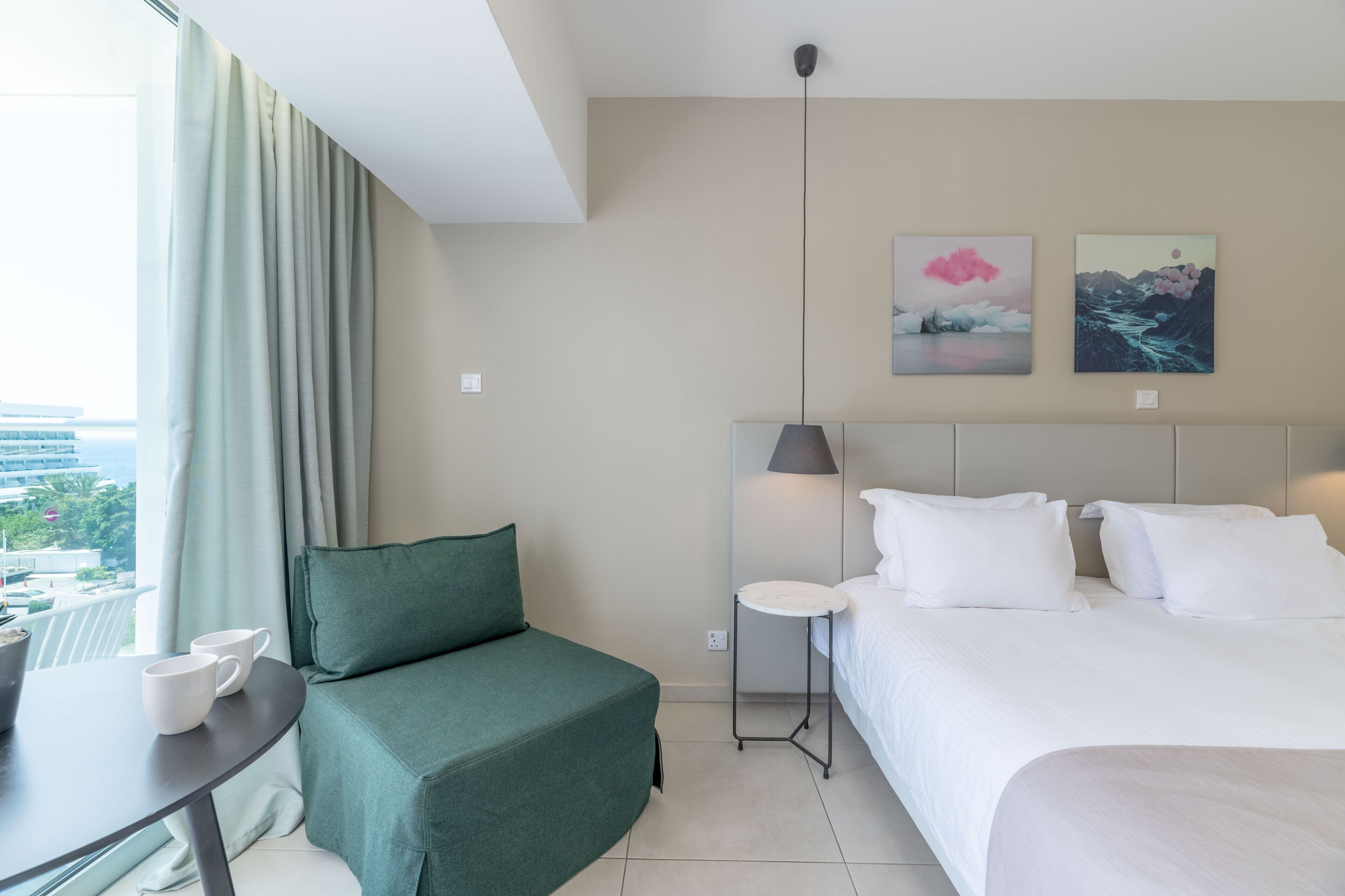 A hotel room with a bed, a green sofa, a small table with two cups, and a view of the city through large windows.
