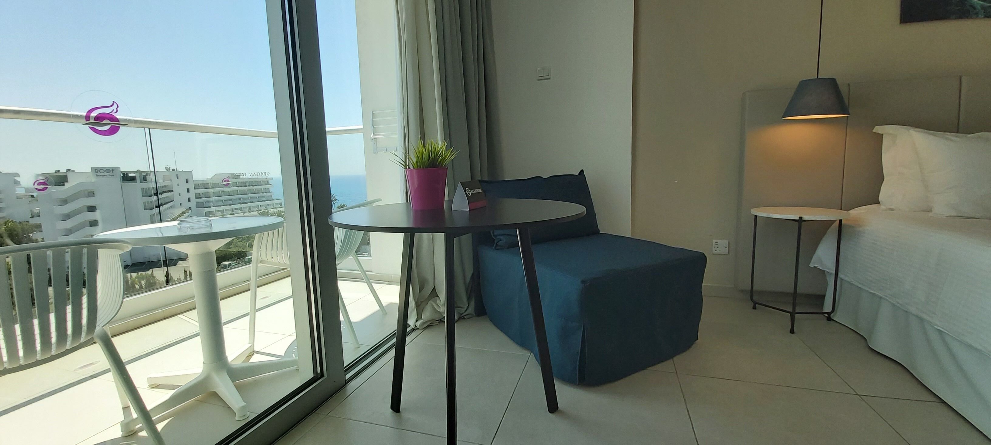 Una habitación de hotel moderna con balcón, una pequeña mesa redonda con una planta en maceta, un sofá azul, una cama con ropa blanca y una lámpara de pie.