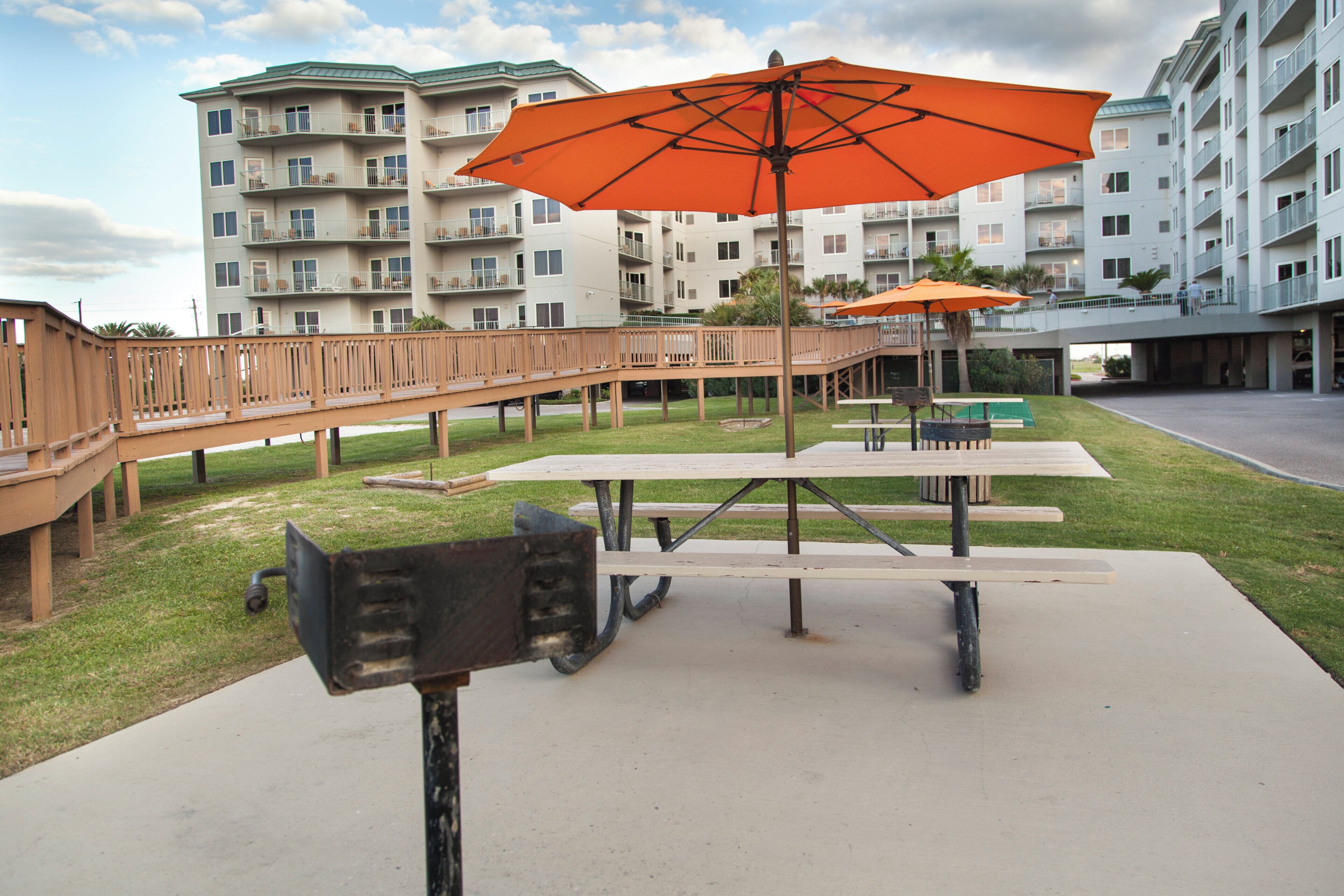 Un espace extérieur aménagé en terrasse, avec des tables de pique-nique et un parasol orange, entouré d’une plateforme en bois et d’un bâtiment doté de balcons.