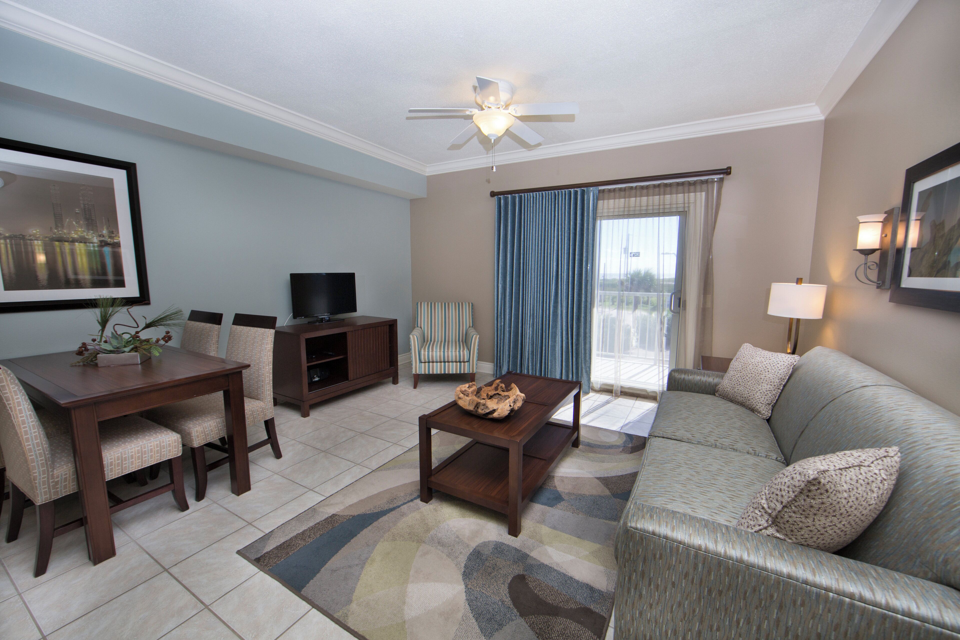A living room with a dining area, a television, a ceiling fan, and a large window with curtains.