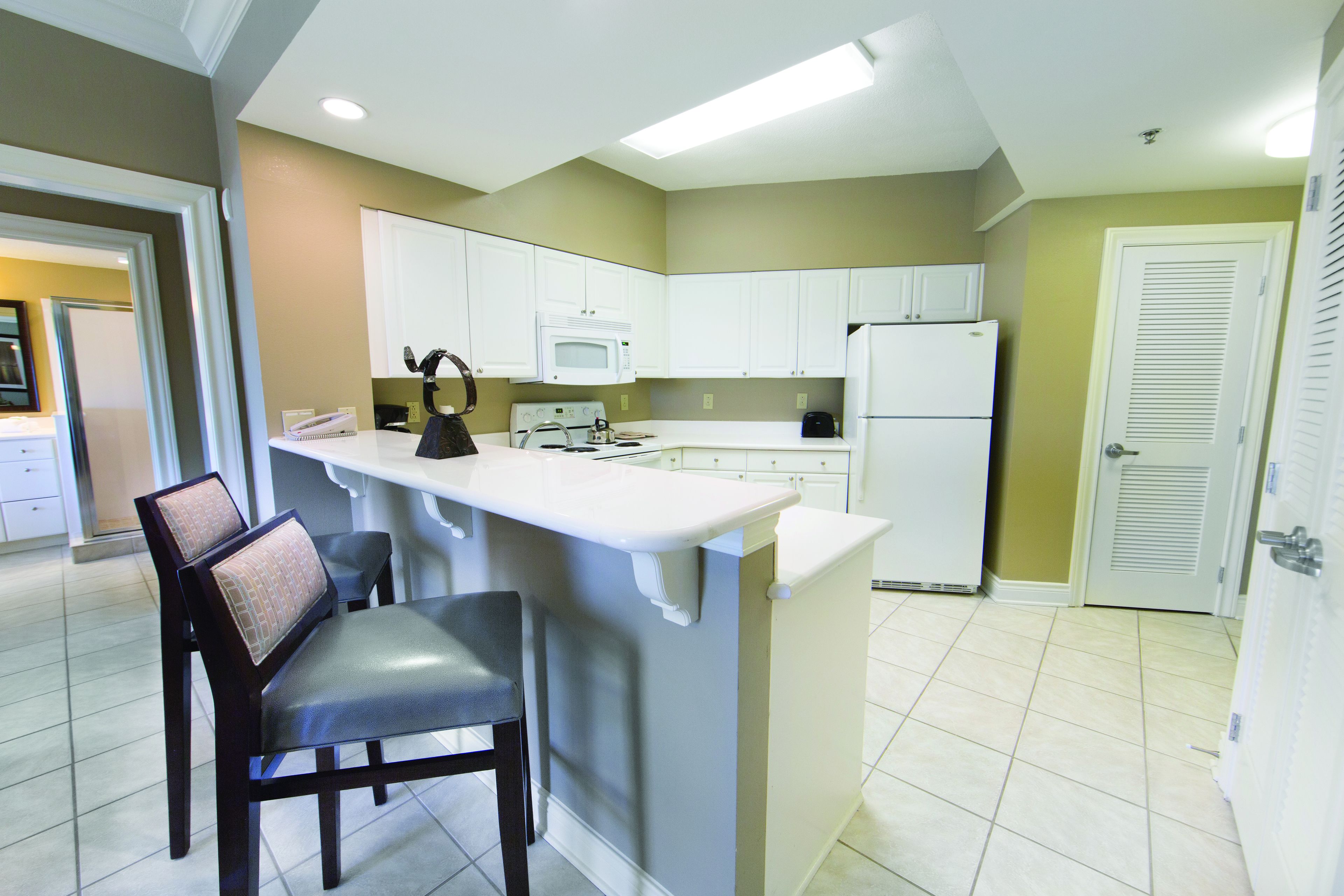 A kitchen with white cabinets, a microwave, a refrigerator, and a bar area with chairs.
