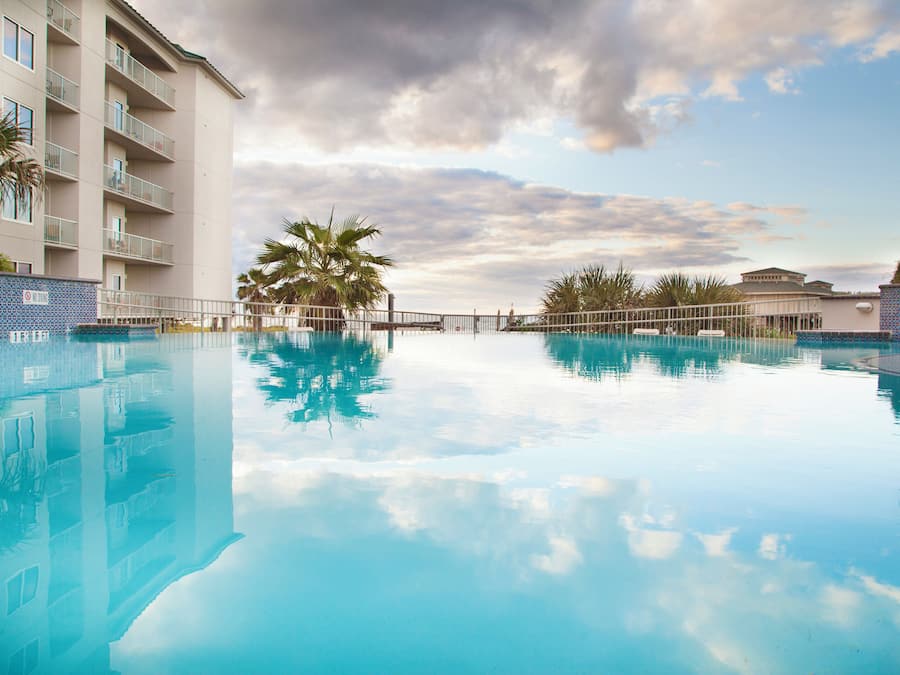 Piscina límpida refletindo o céu e palmeiras, rodeada por um edifício com varandas.