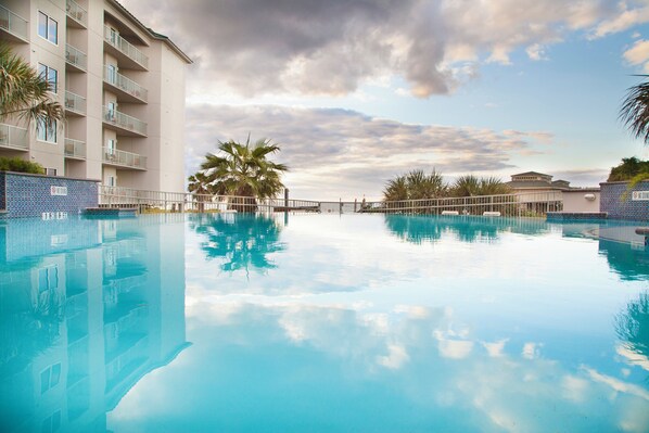 A clear swimming pool with a reflection of the sky and palm trees, surrounded by a building with balconies.