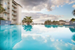 En klar pool där himlen och palmerna speglas, omgiven av en byggnad med balkonger.