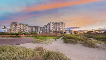 A beachfront hotel with multiple balconies, a sandy beach in the foreground, and a sunset sky.