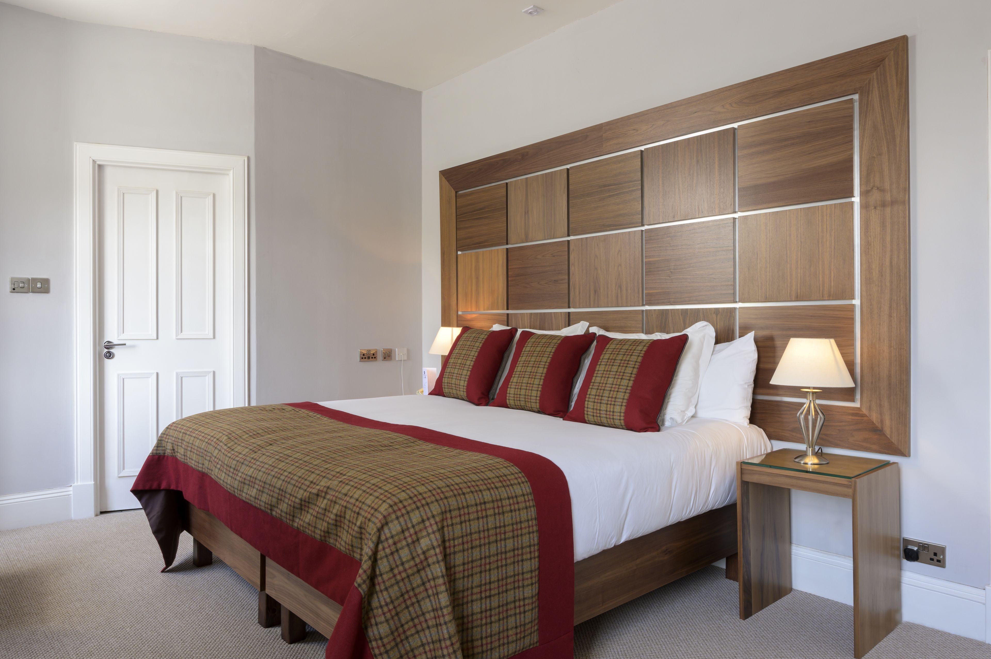 A neatly made bed with a wooden headboard, a bedside table with a lamp, and a door to the left.