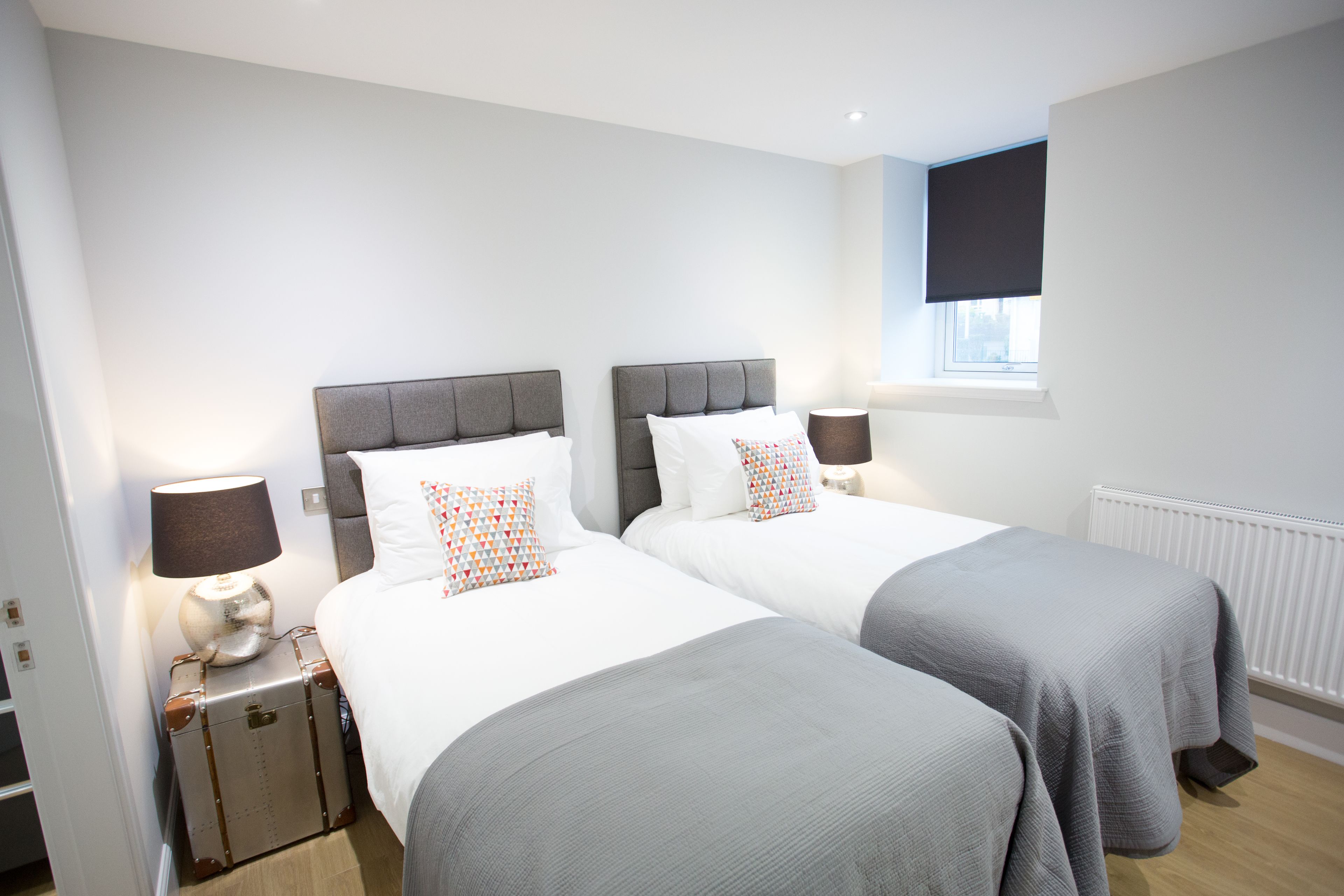 A hotel room with two beds, each with a grey headboard and white bedding. There is a lamp on the nightstand beside each bed, and a suitcase is placed on the floor next to one of the beds.