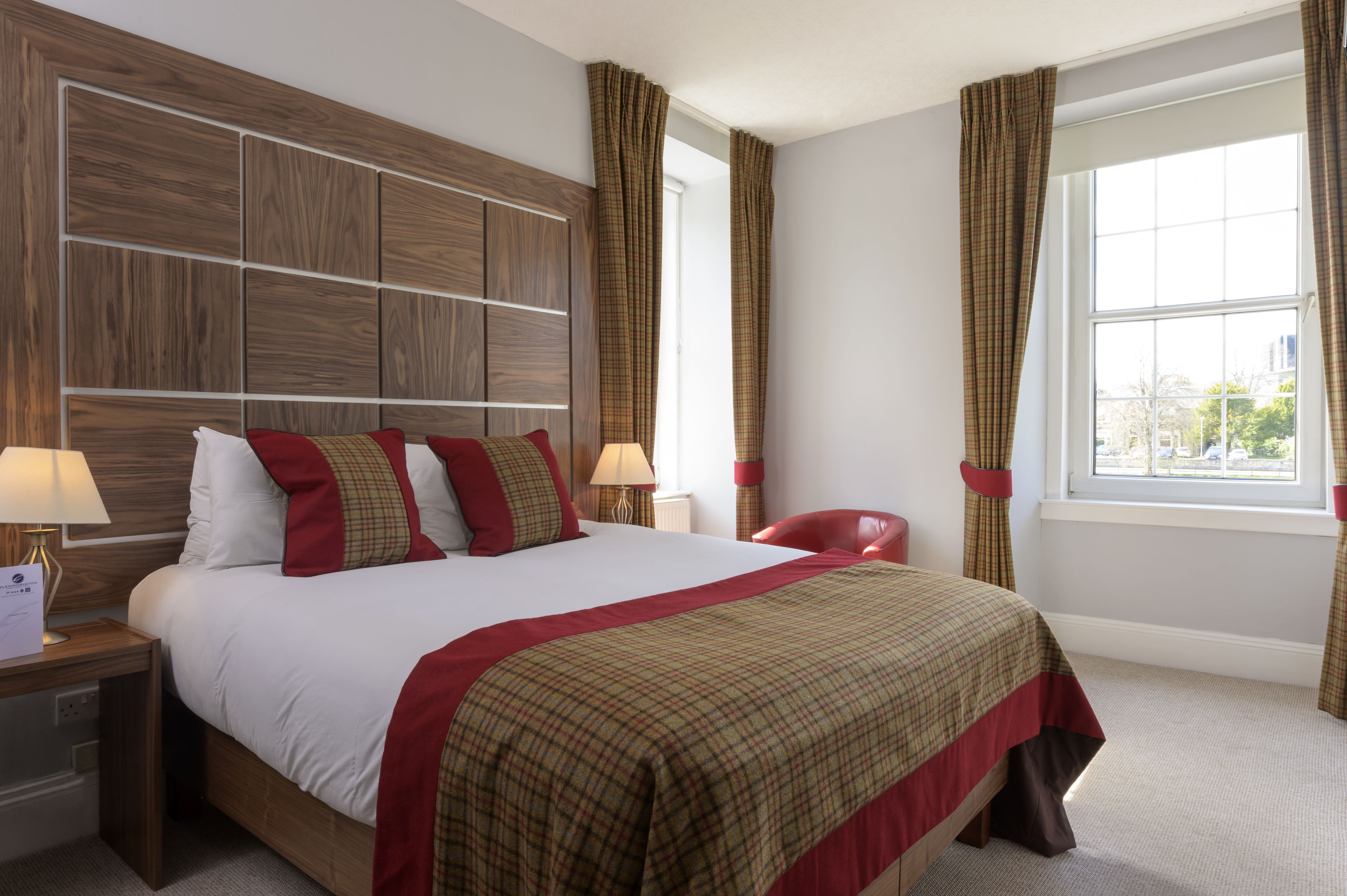 A hotel room with a large bed, wooden headboard, two bedside tables with lamps, a red chair, and a window with curtains.