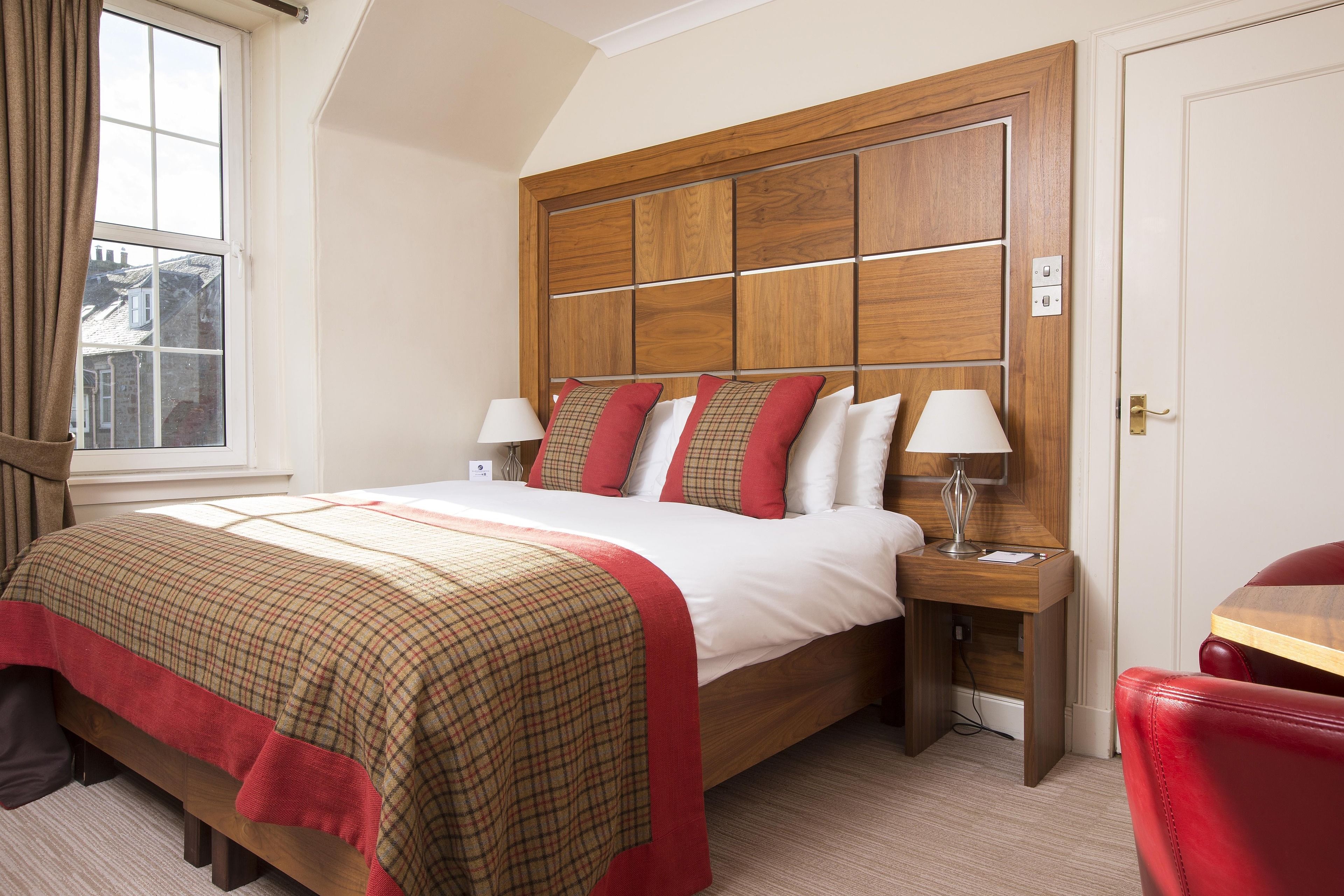 Ein Hotelzimmer mit einem großen Bett, einem Holzkopfteil, zwei Nachttischen mit Lampen, einem roten Sessel und einem Fenster mit Vorhängen.