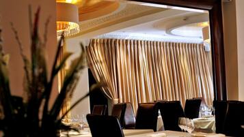 A dining area with a table set for two, a chandelier, and a view through a window with curtains.