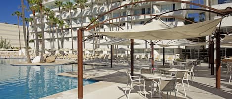 A poolside area with lounge chairs and umbrellas, a pool, and a building with balconies and palm trees.