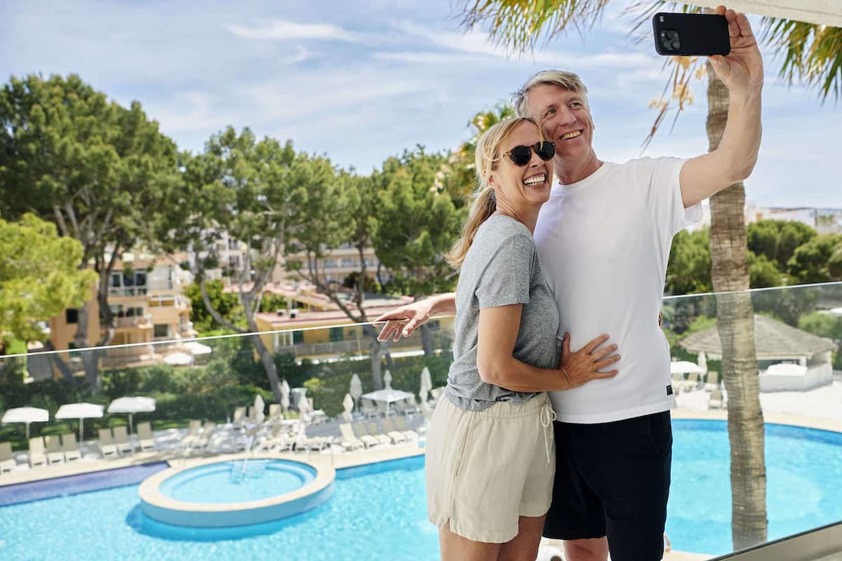 Een koppel maakt een selfie bij een zwembad met uitzicht op een resort, inclusief een zwembad en ligstoelen.