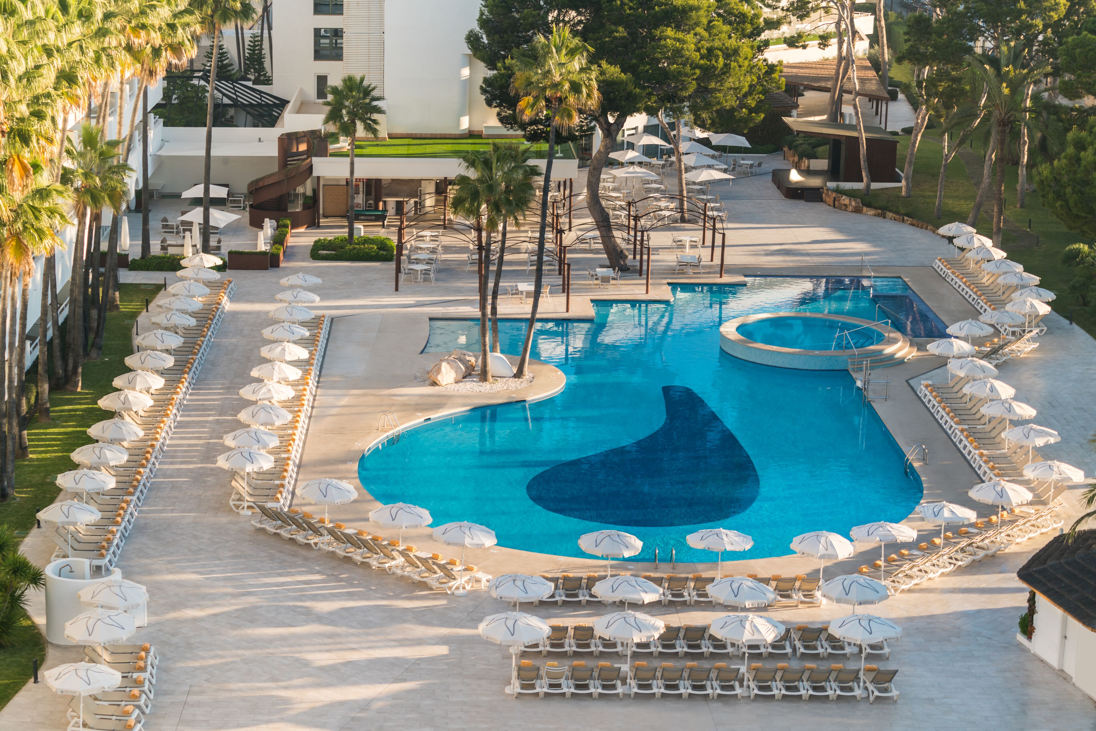 Piscina de un resort con tumbonas y sombrillas, rodeada de palmeras y un edificio de diseño moderno.