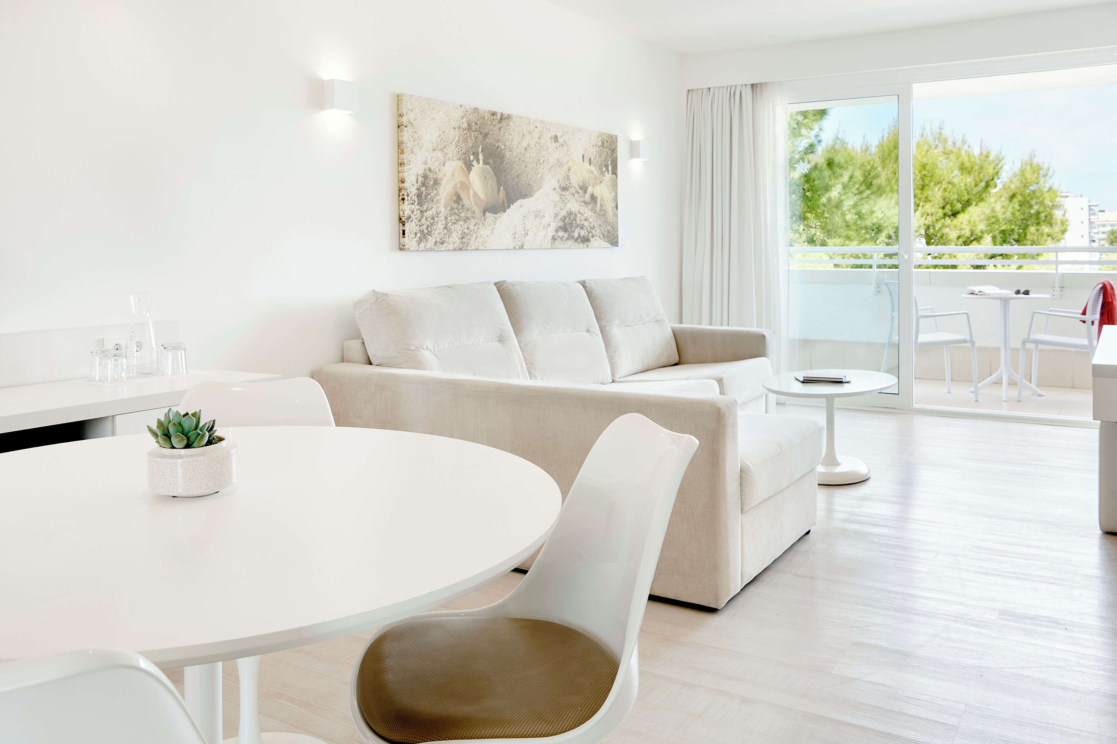 Un salon moderne avec un canapé blanc, une table de salle à manger ronde et un balcon offrant une vue sur les arbres.
