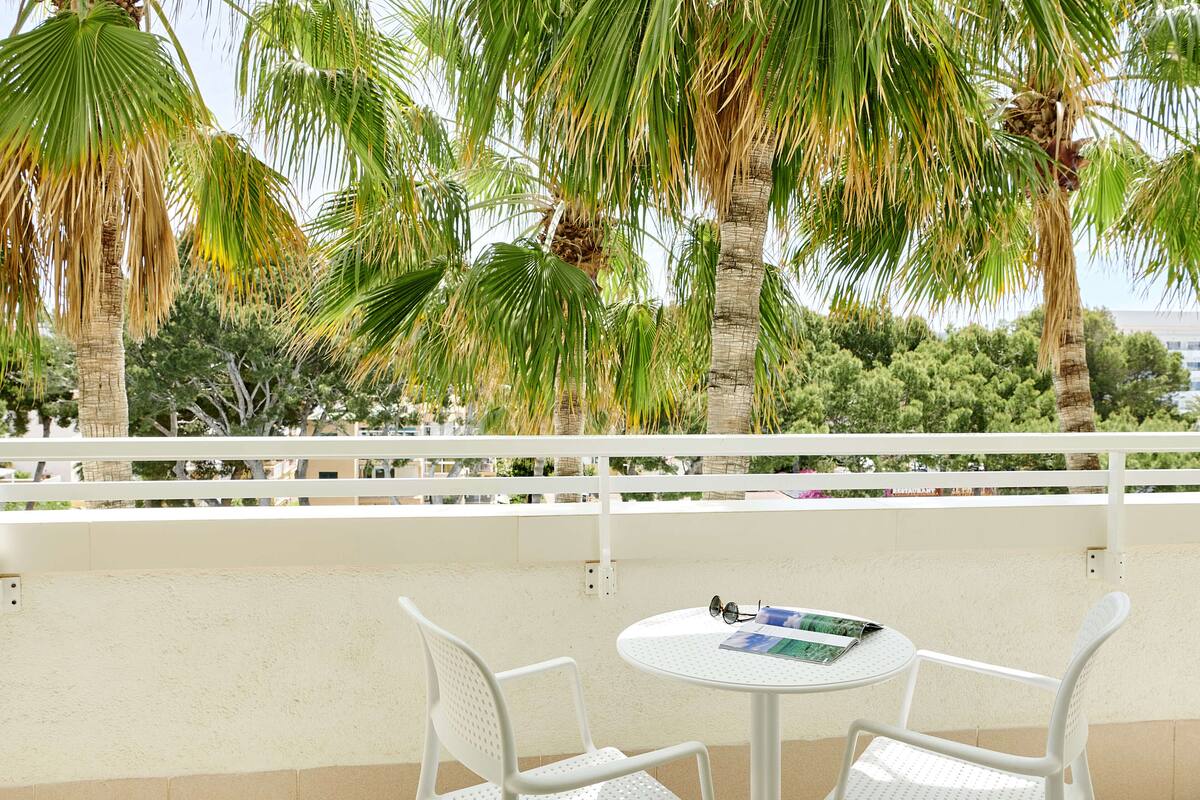 Een balkon met witte stoelen en een tafel, palmbomen en een uitzicht op gebouwen.