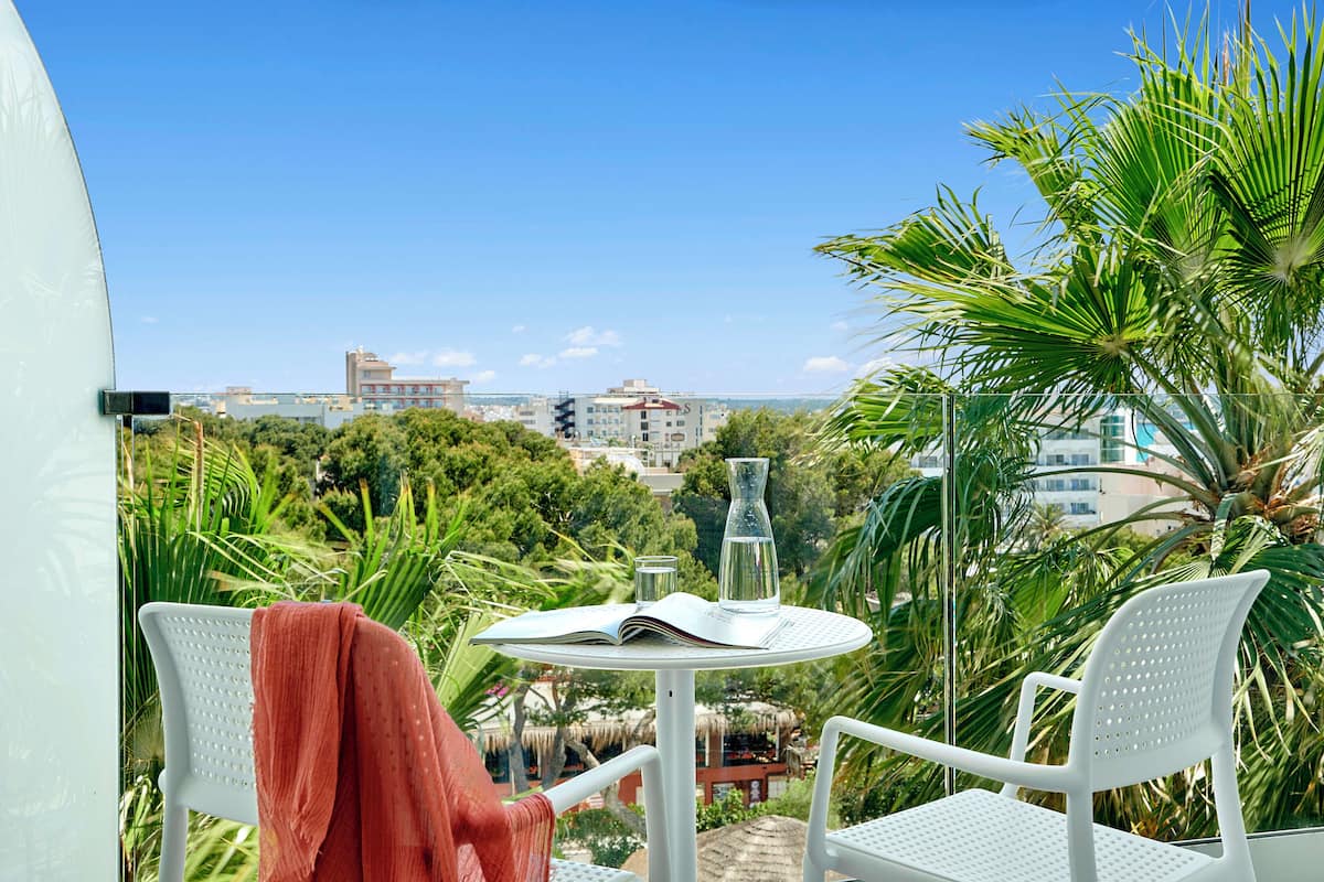 Ein Balkon mit weißen Stühlen, einem Tisch mit Buch und Glasvase sowie Blick auf Gebäude und Bäume.