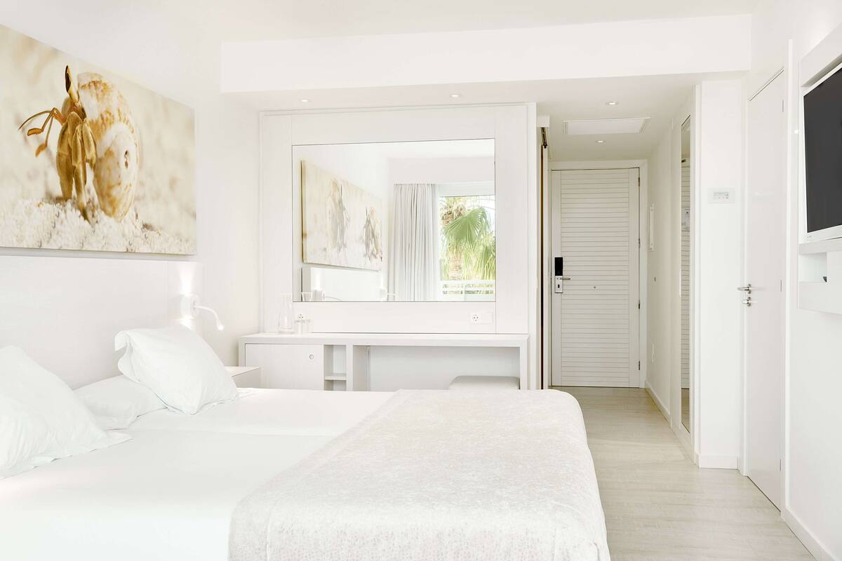 Een moderne slaapkamer met een groot bed, een witte ladekast, een spiegel en een televisie.