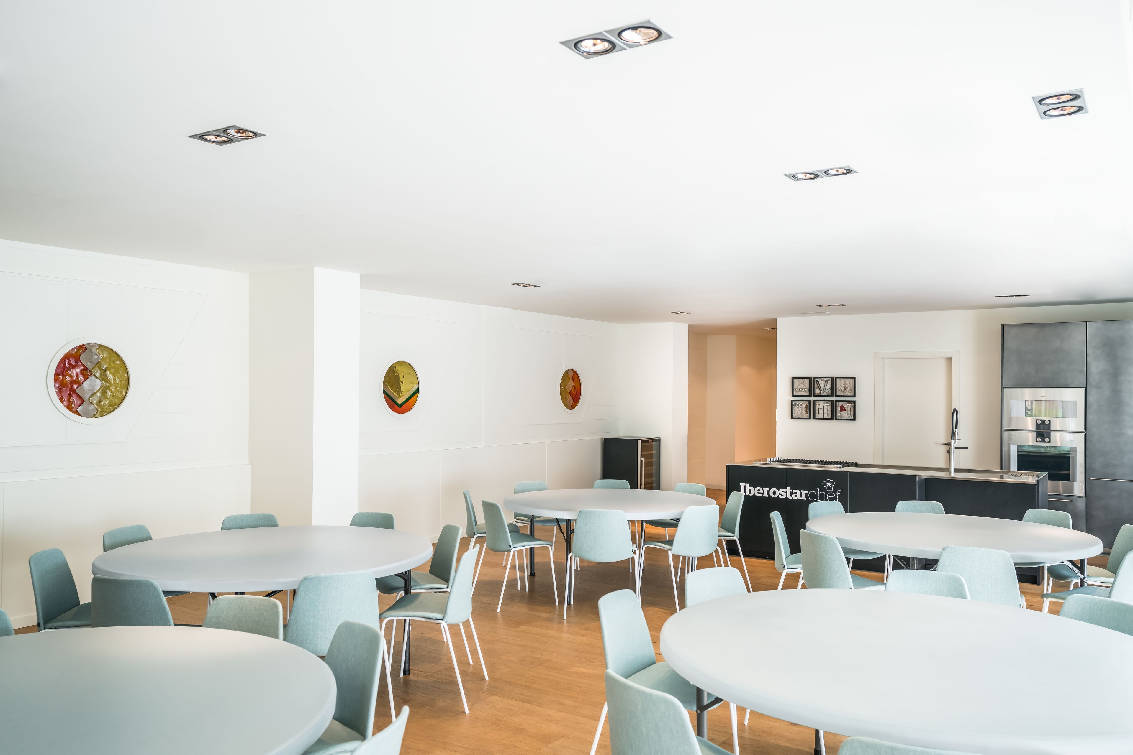 Una sala de conferencias moderna con mesas redondas y sillas azul claro, una zona de cocina con un refrigerador de marca y decoración en las paredes.