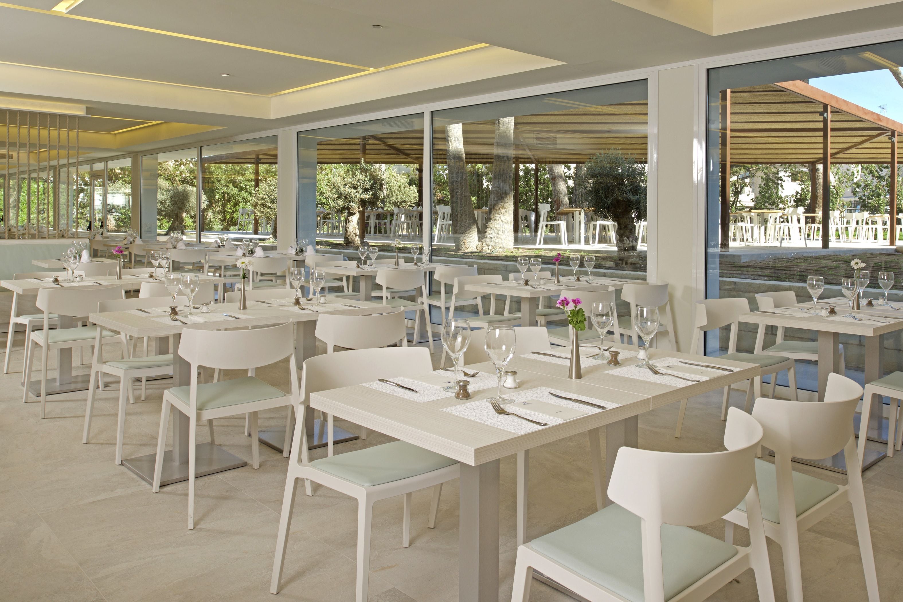 A restaurant with white tables and chairs, large windows, and a view of the outdoors.