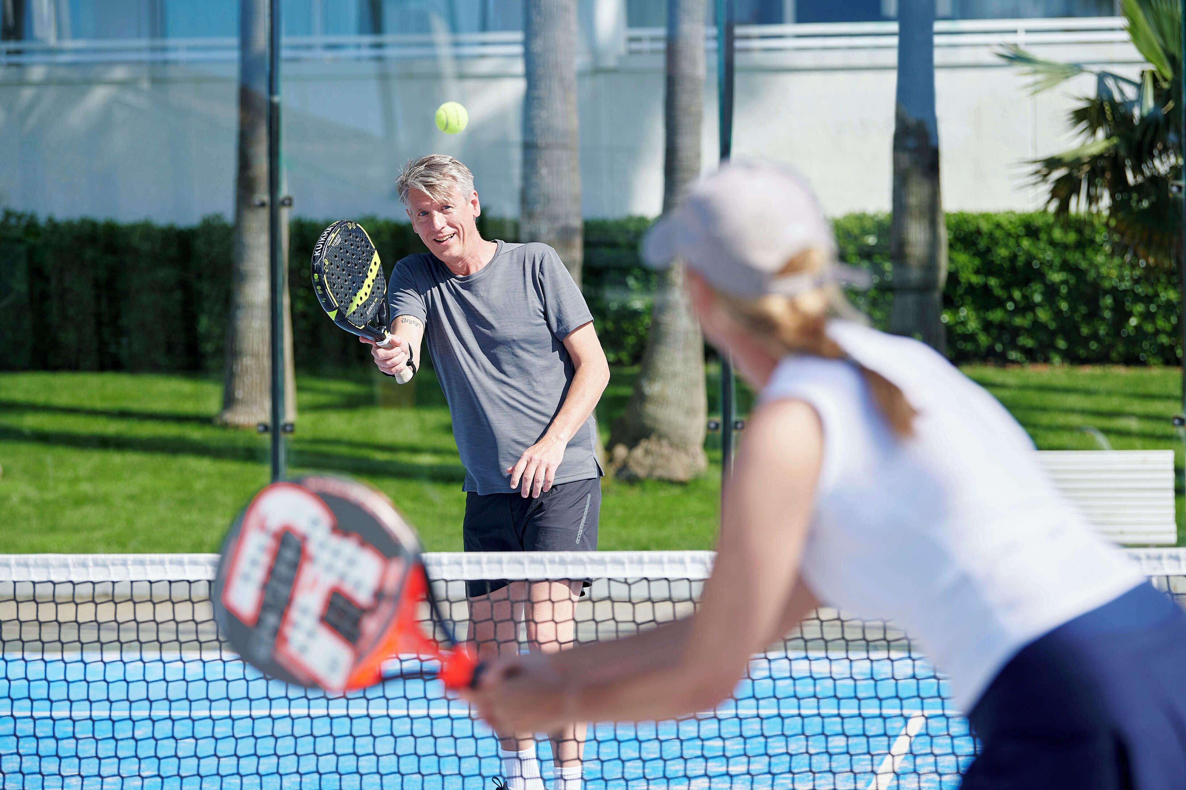 En tennisbane med et net, en tennisbold i luften, og to personer der spiller.