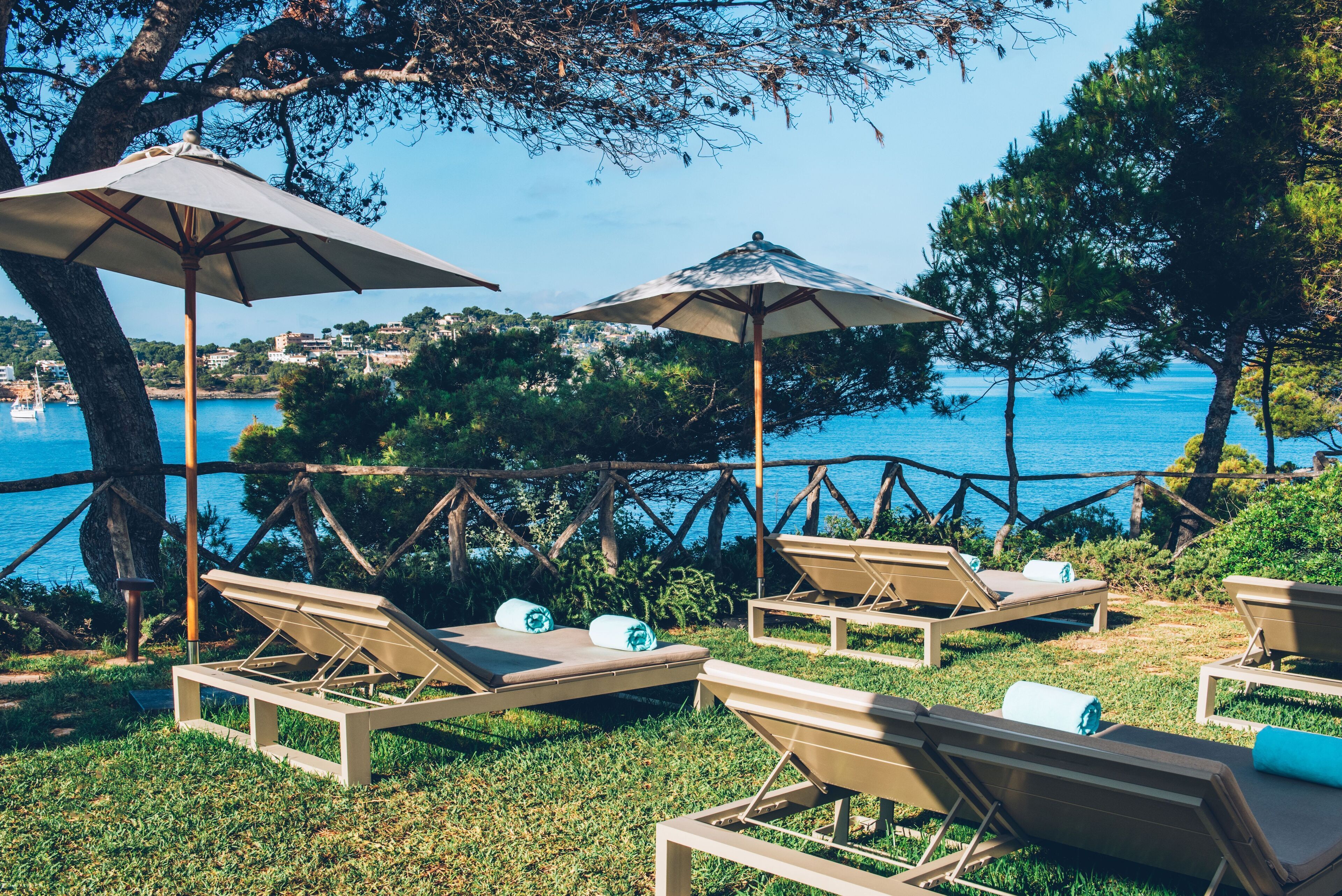 Sillas de lounge al aire libre con sombrillas, un área verde y vistas al mar y edificios al fondo.