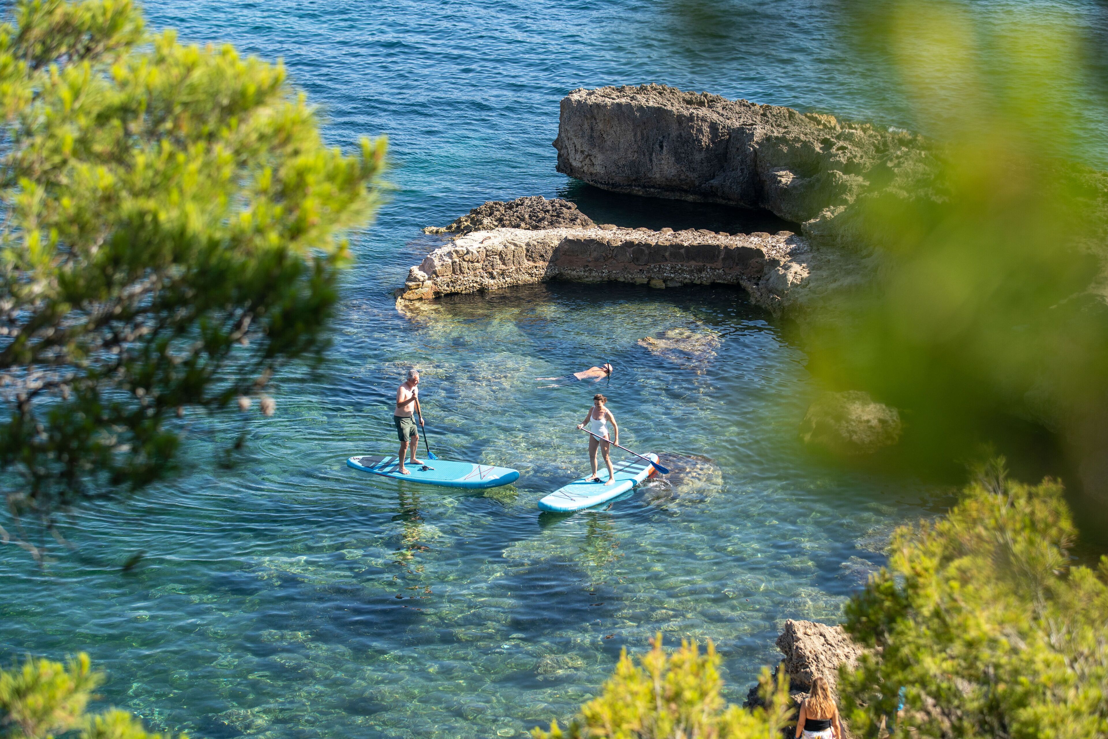 Two people paddleboarding on clear water with rocky formations in the background.