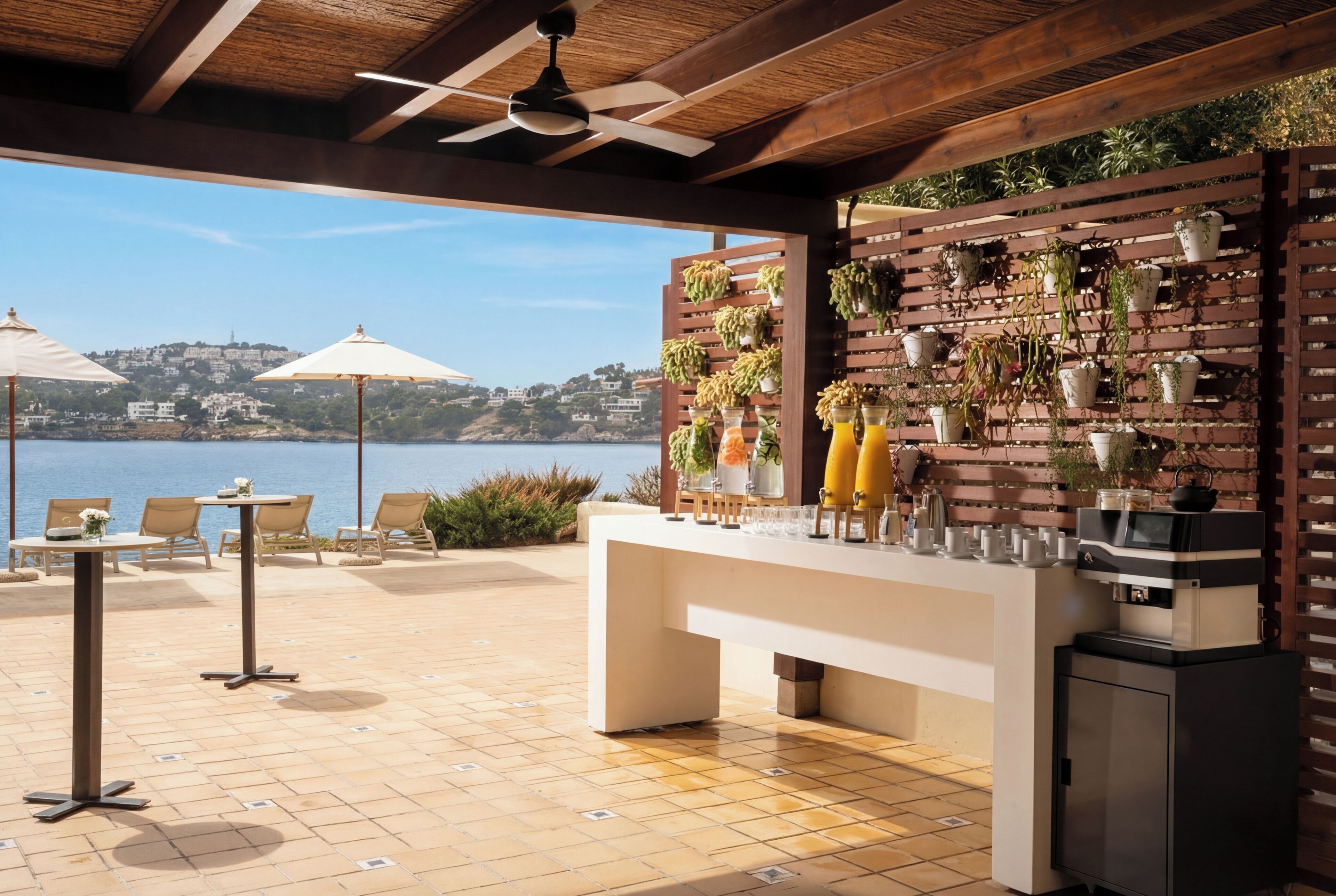 An outdoor bar area with a view of the sea, a wooden wall with potted plants, and a ceiling fan.