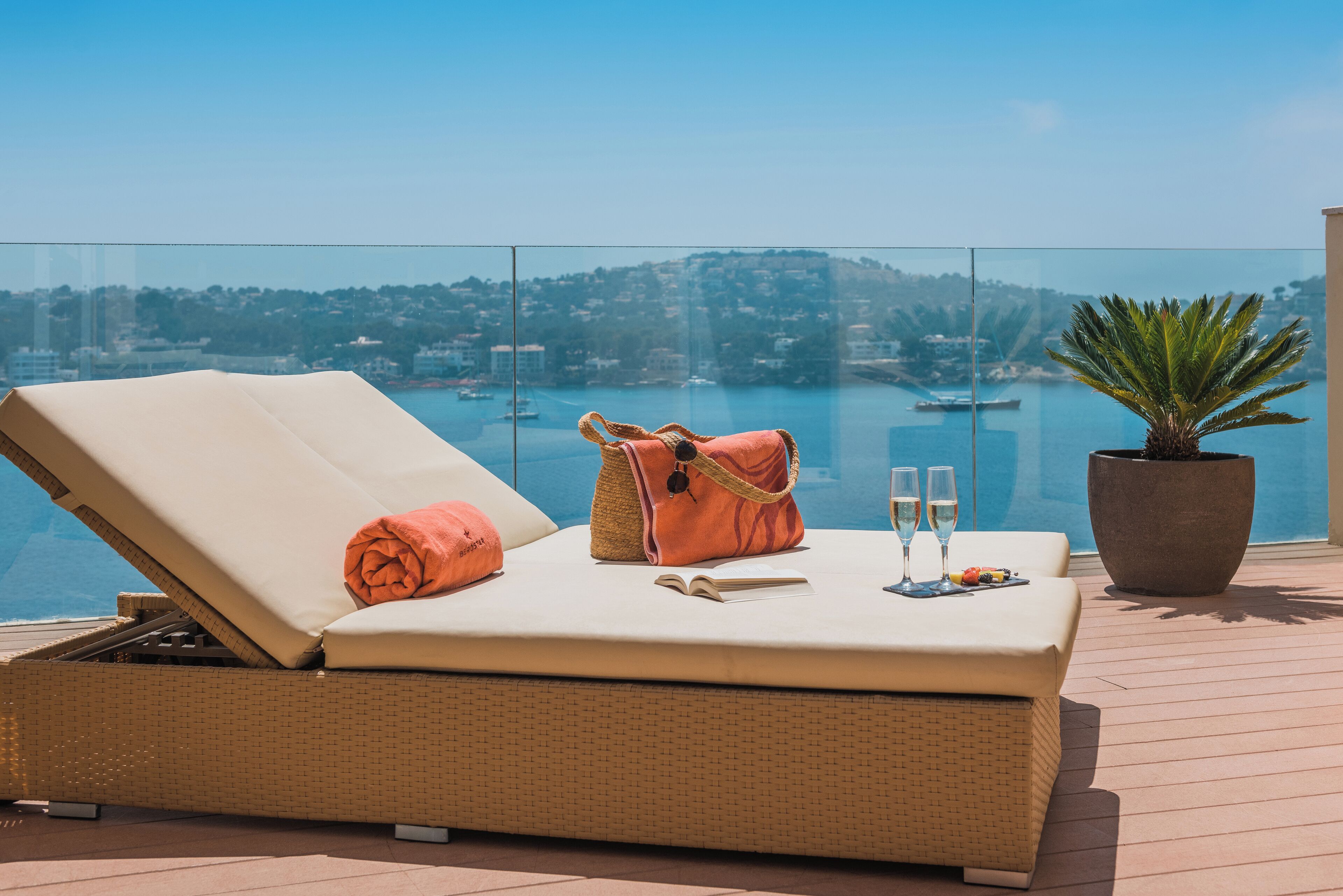 Une chaise longue sur laquelle est posée une serviette, un sac et deux coupes de champagne, sur un balcon offrant une vue sur une étendue d’eau et des bâtiments.