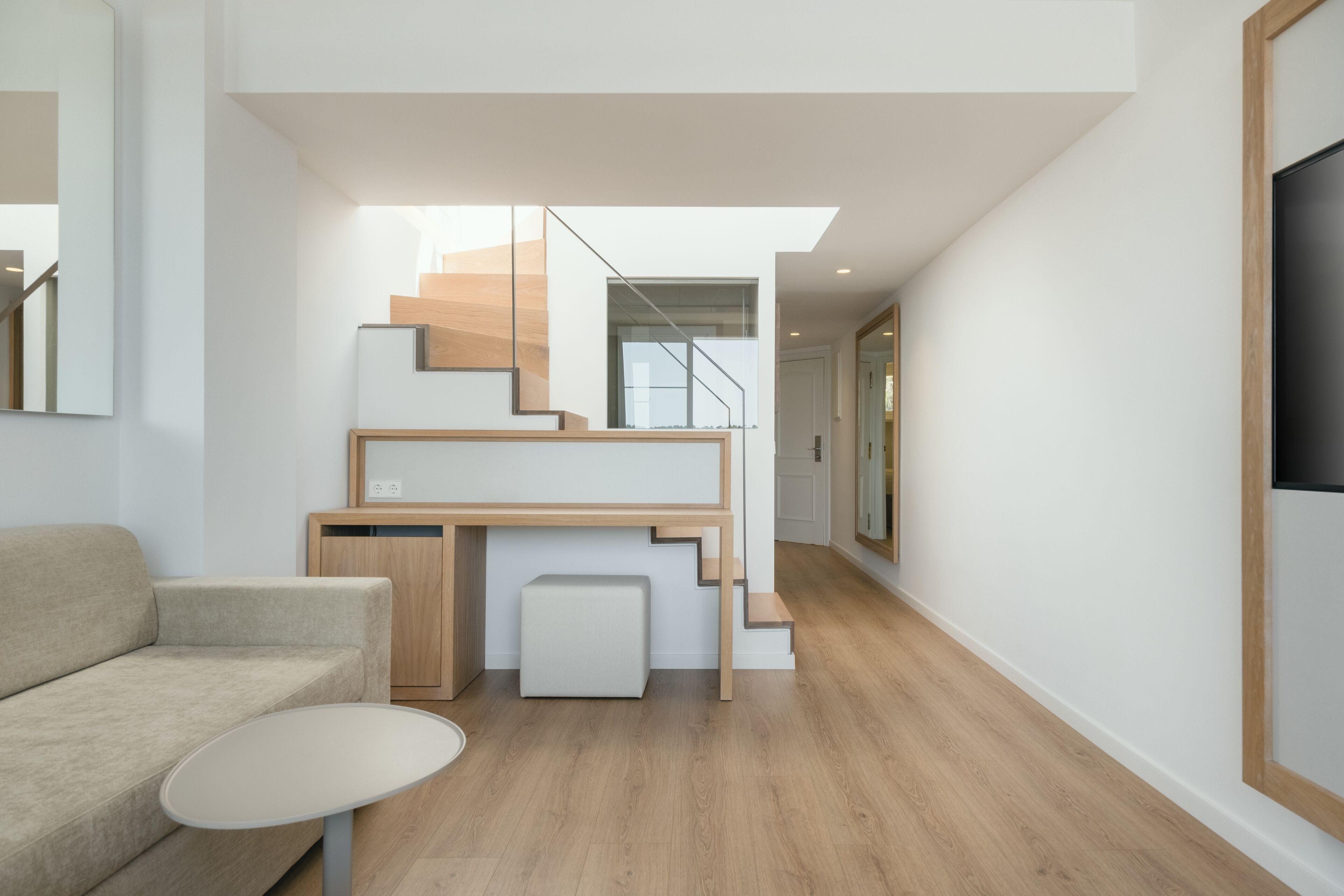 Un interior moderno con un sofá, una mesita redonda, un escritorio de madera con una silla, y una escalera que sube a un nivel superior.