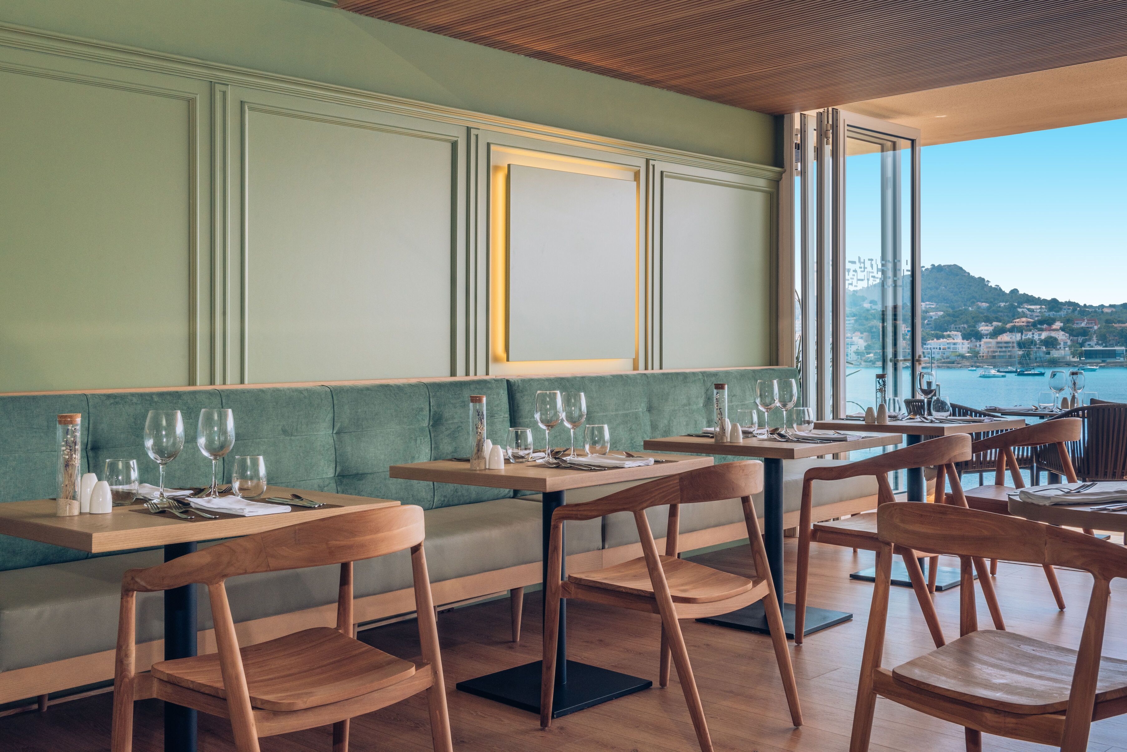 A restaurant with wooden tables and chairs, a green cushioned bench, and a view of the sea.