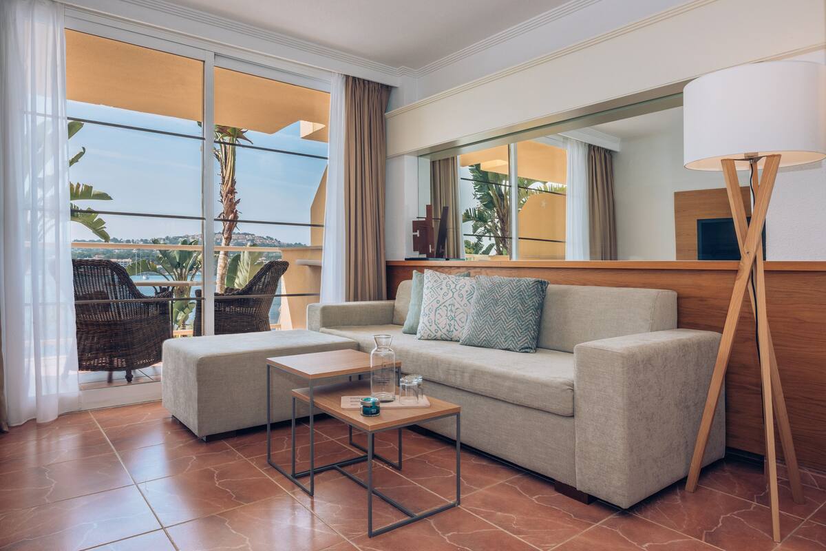 Een moderne woonkamer met een bank, salontafel en vloerlamp. Er is een balkon met rieten meubilair en uitzicht op het landschap.