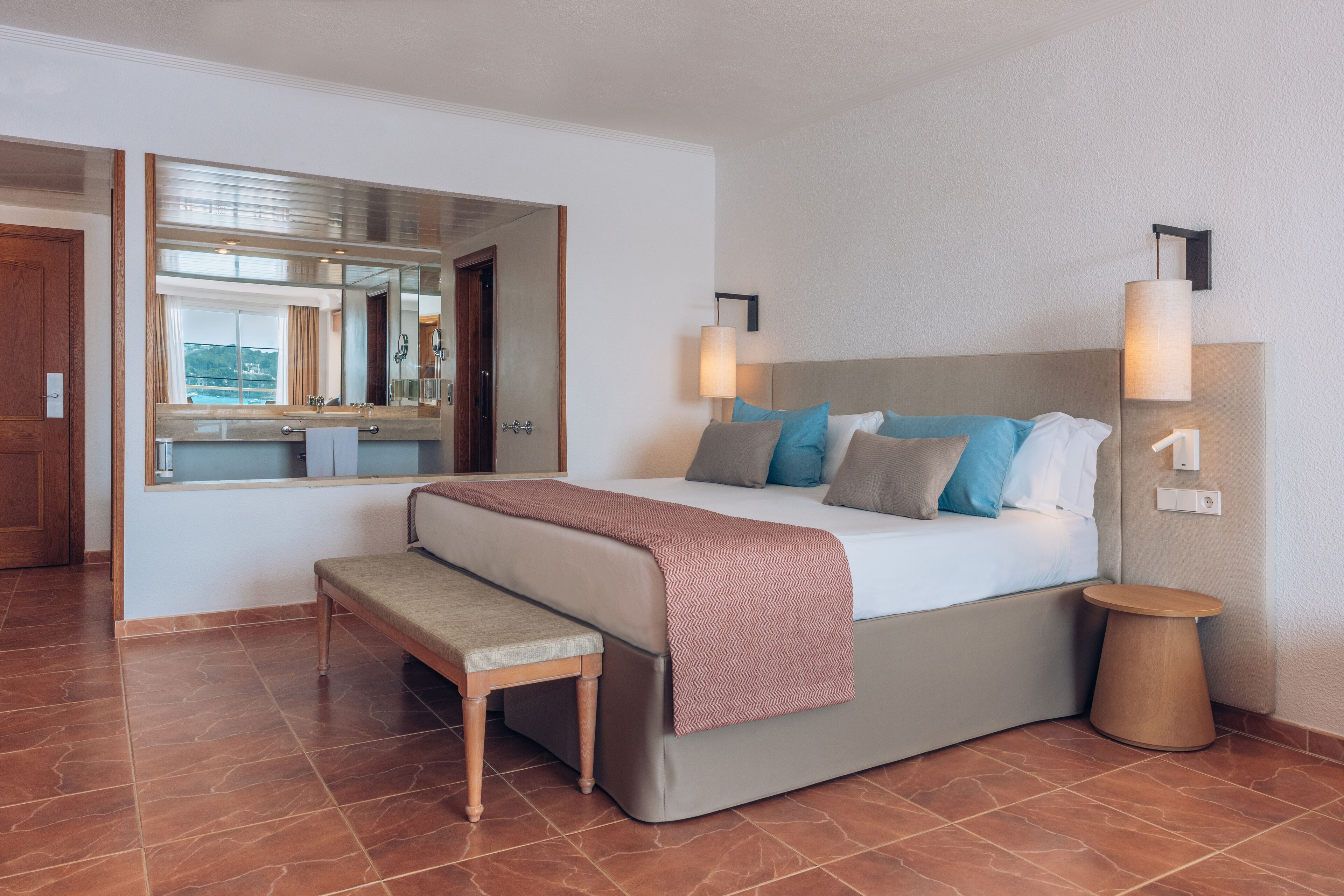 Habitación de hotel moderna con una cama grande, un banco, una mesita pequeña y un espejo que refleja la habitación.