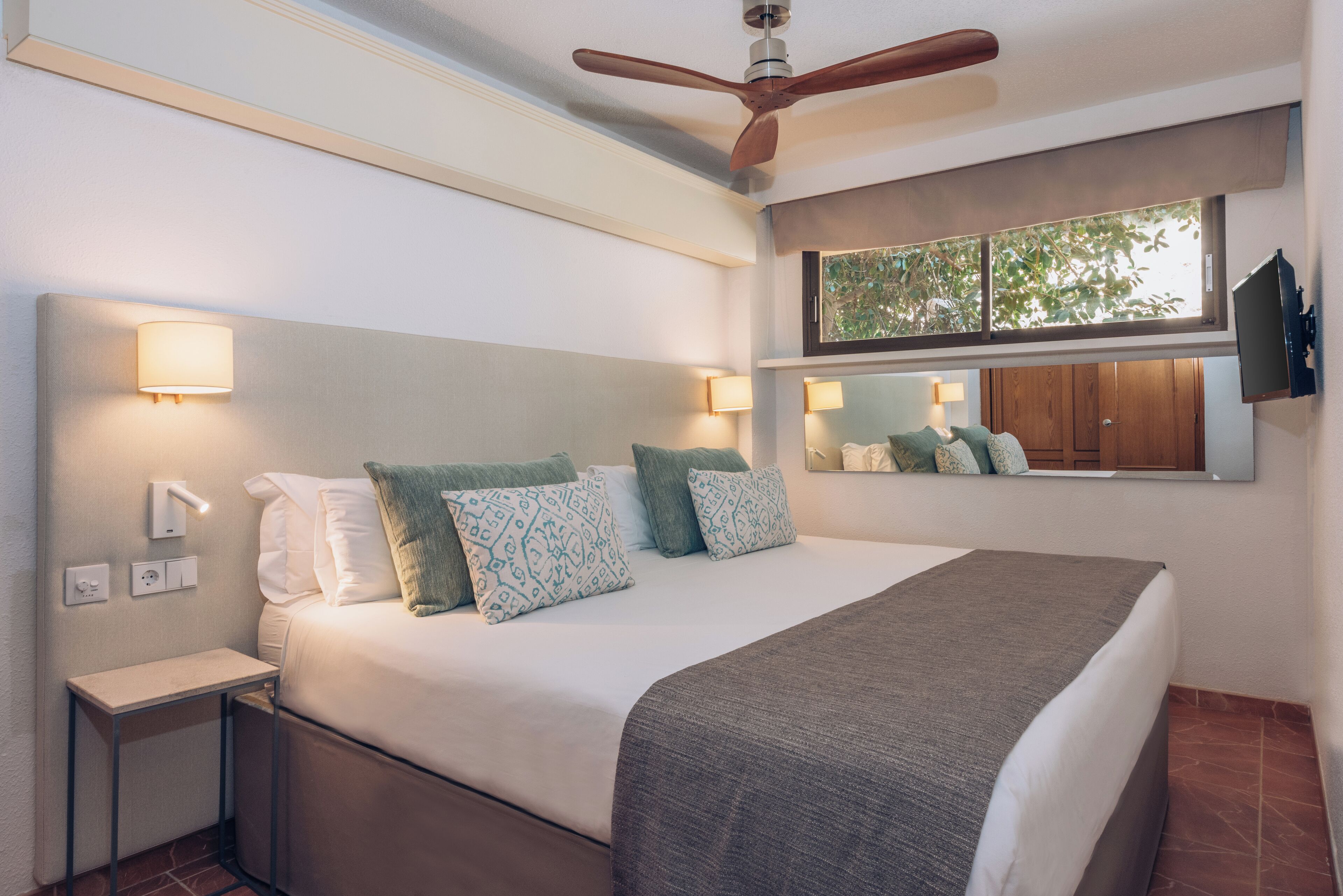 A hotel room with a bed, bedside lamps, a ceiling fan, a window with a view, and a mounted TV.