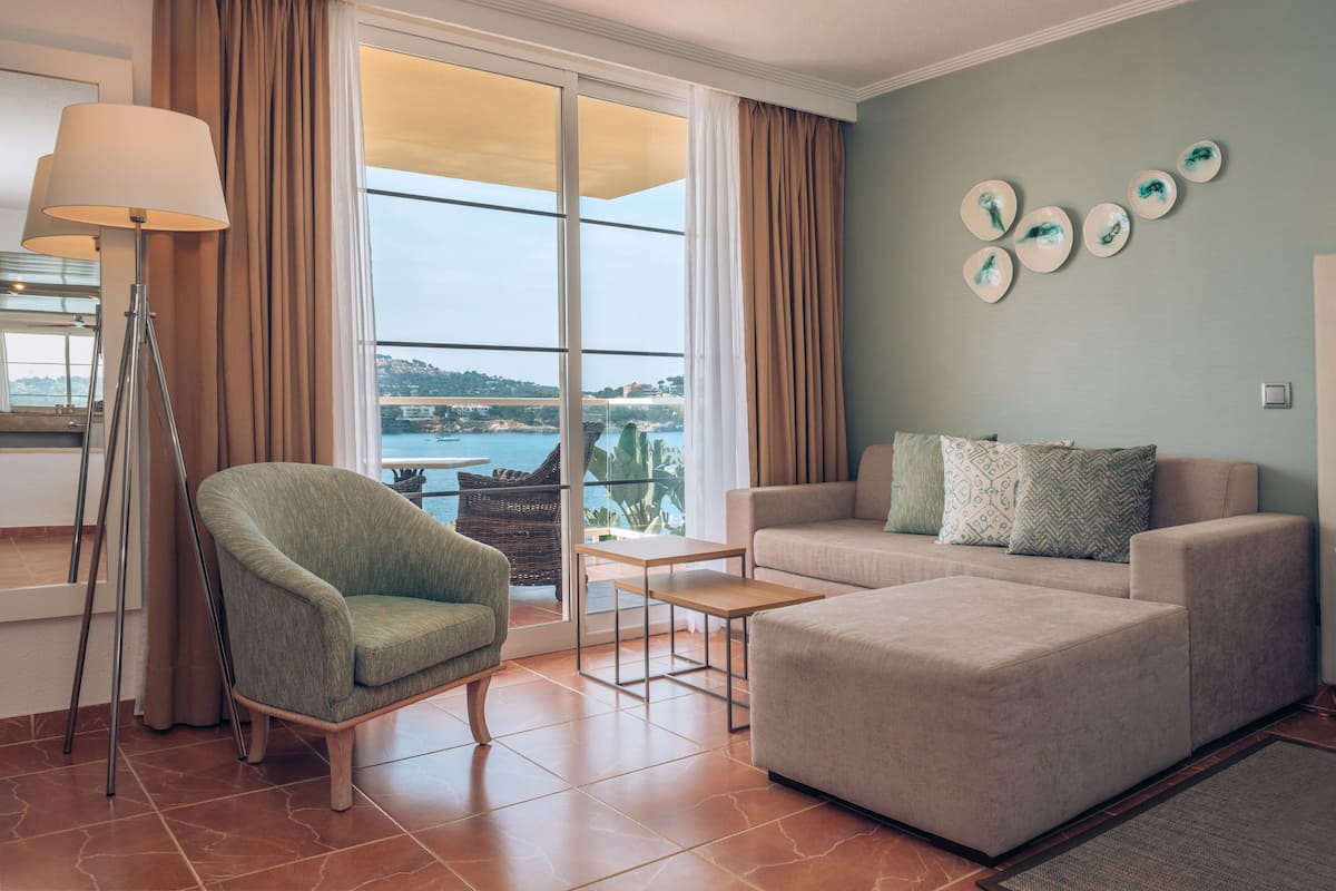 Een moderne woonkamer met een bank, fauteuil en een vloerlamp. Een groot raam biedt uitzicht op zee en een verafgelegen landschap.