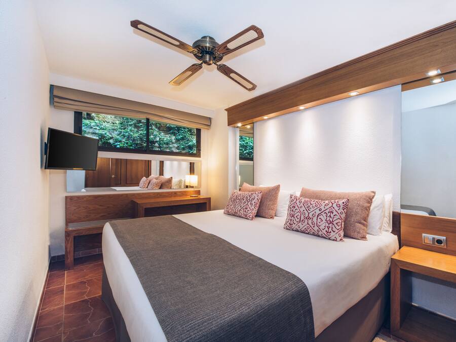 A modern hotel room with a large bed, a ceiling fan, a TV, and a window with a view of trees.