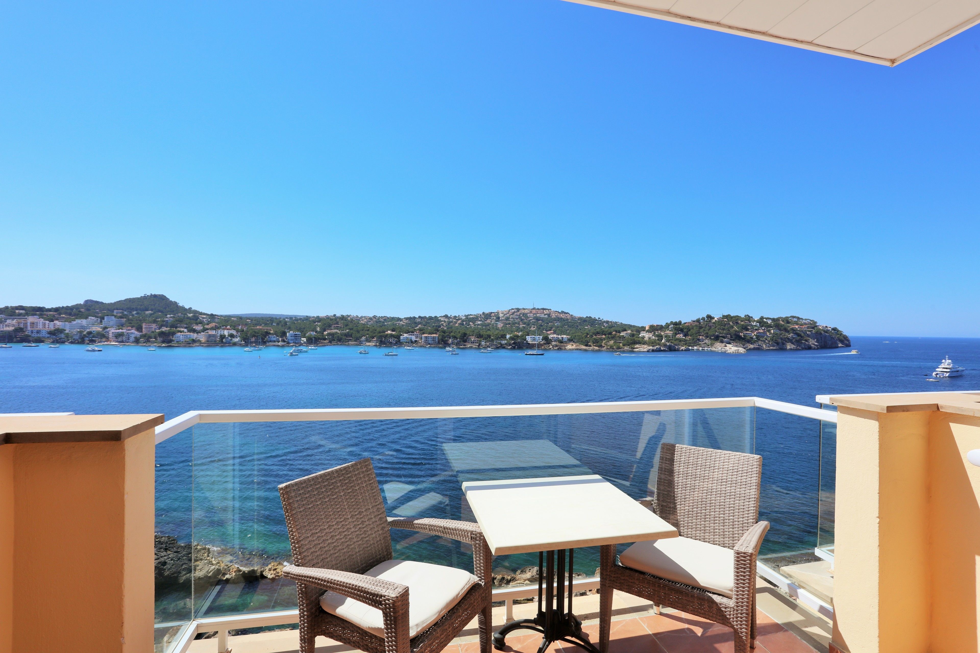 Ein Balkon mit Korbstühlen und einem Tisch, der Blick auf eine Küstenlandschaft mit Booten und Gebäuden bietet.