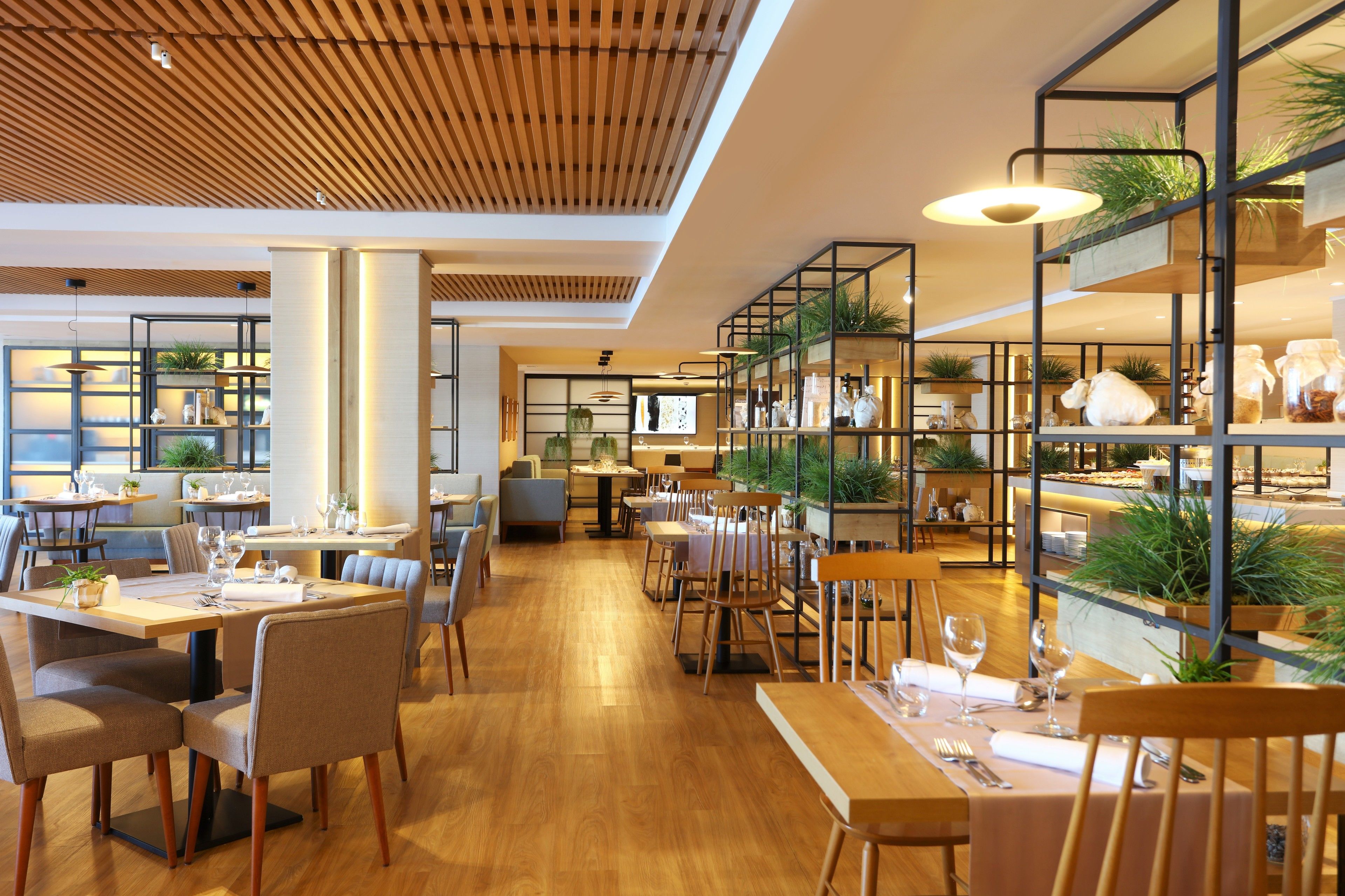A modern restaurant with wooden flooring, multiple dining areas, and shelves with plants and kitchenware.