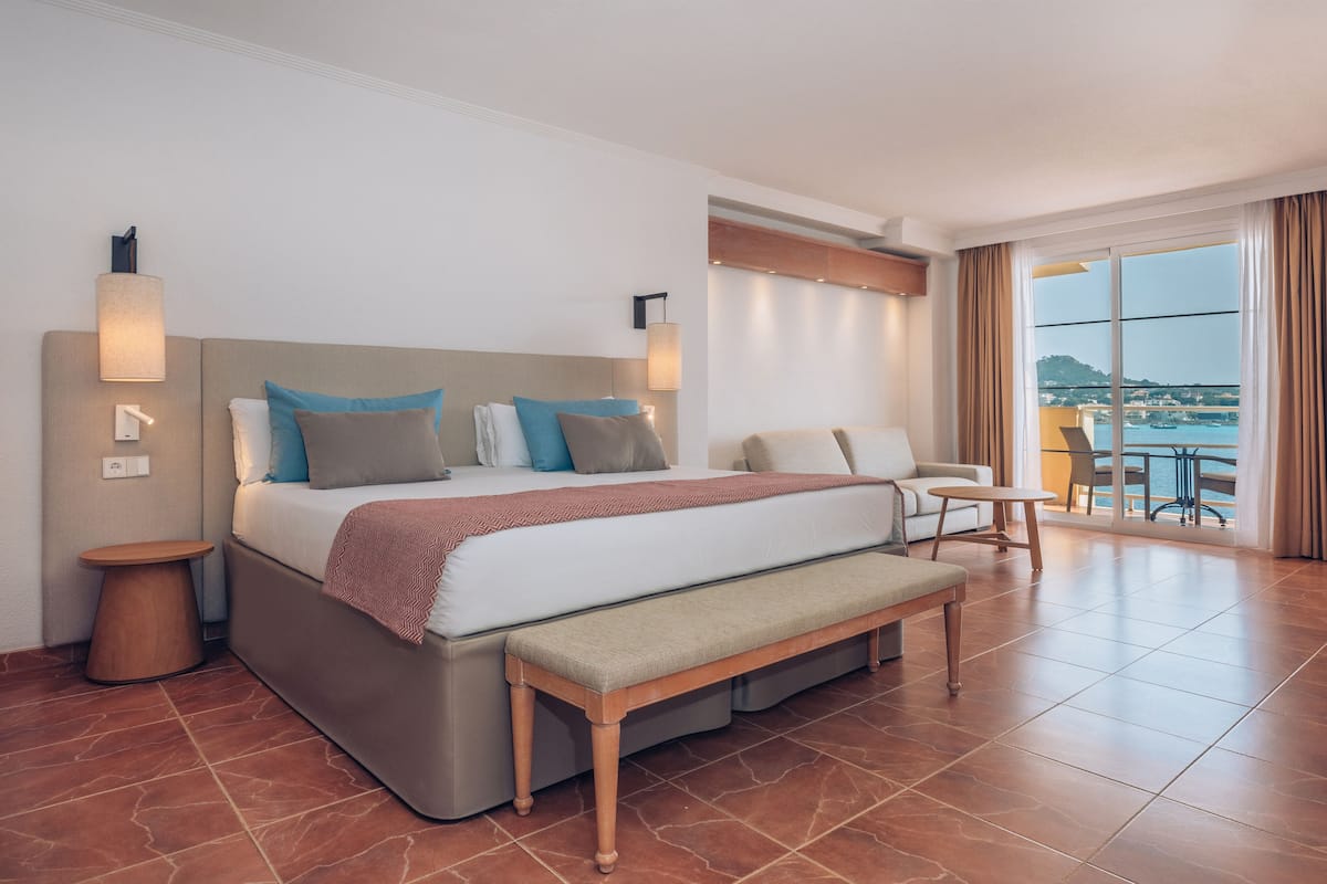 Een moderne hotelkamer met een groot bed, een bankje, een tafeltje en een balkon met uitzicht op zee.