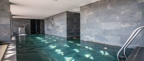 A modern indoor swimming pool with clear water, surrounded by dark stone walls and a metal ladder.