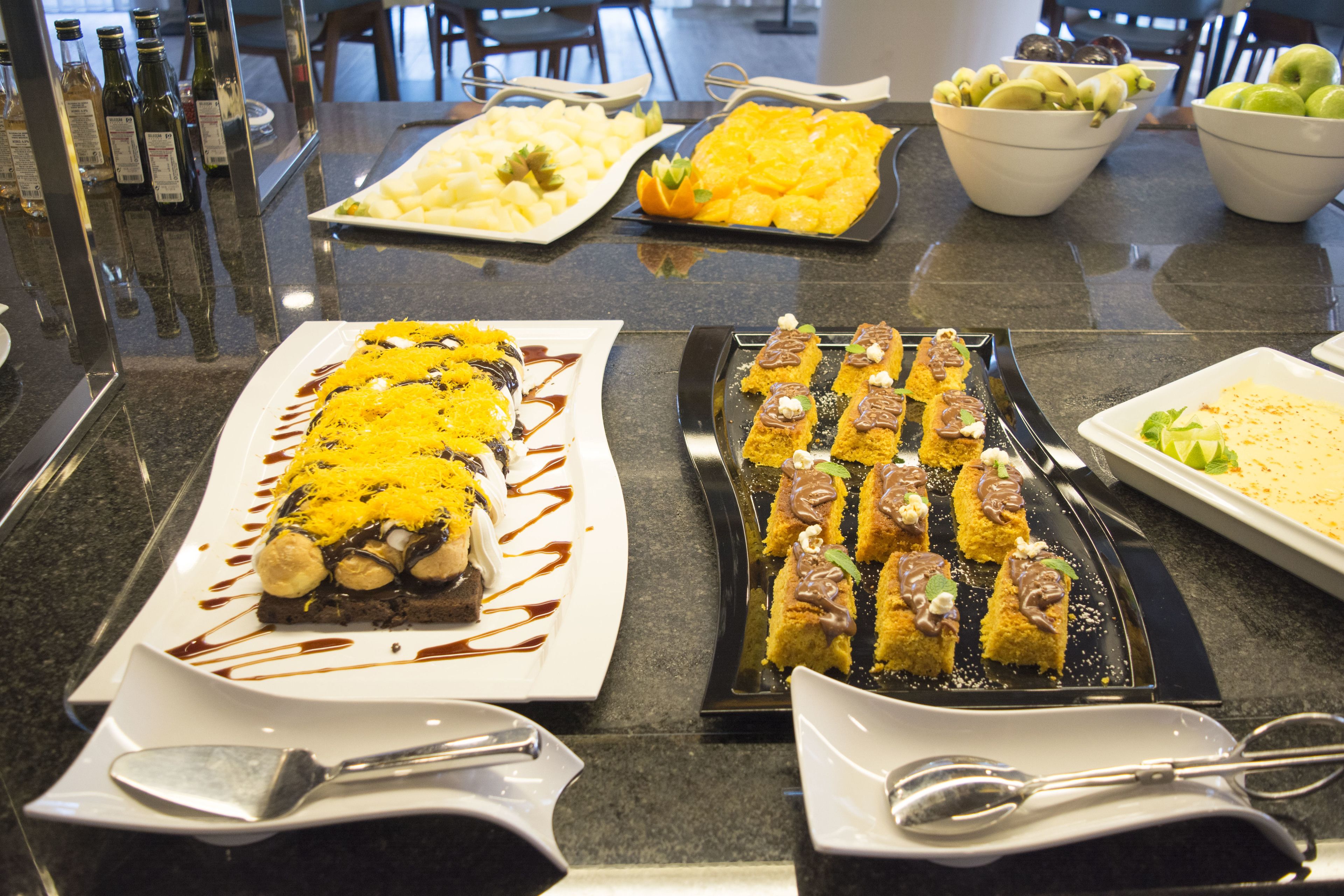 Una mesa de buffet con diversos platos, incluyendo una fuente larga con comida amarilla, una bandeja de pequeños postres y cuencos con frutas verdes y amarillas.