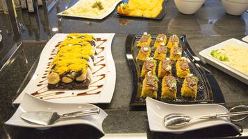 Una mesa de buffet con diversos platos, incluyendo una fuente larga con comida amarilla, una bandeja de pequeños postres y cuencos con frutas verdes y amarillas.