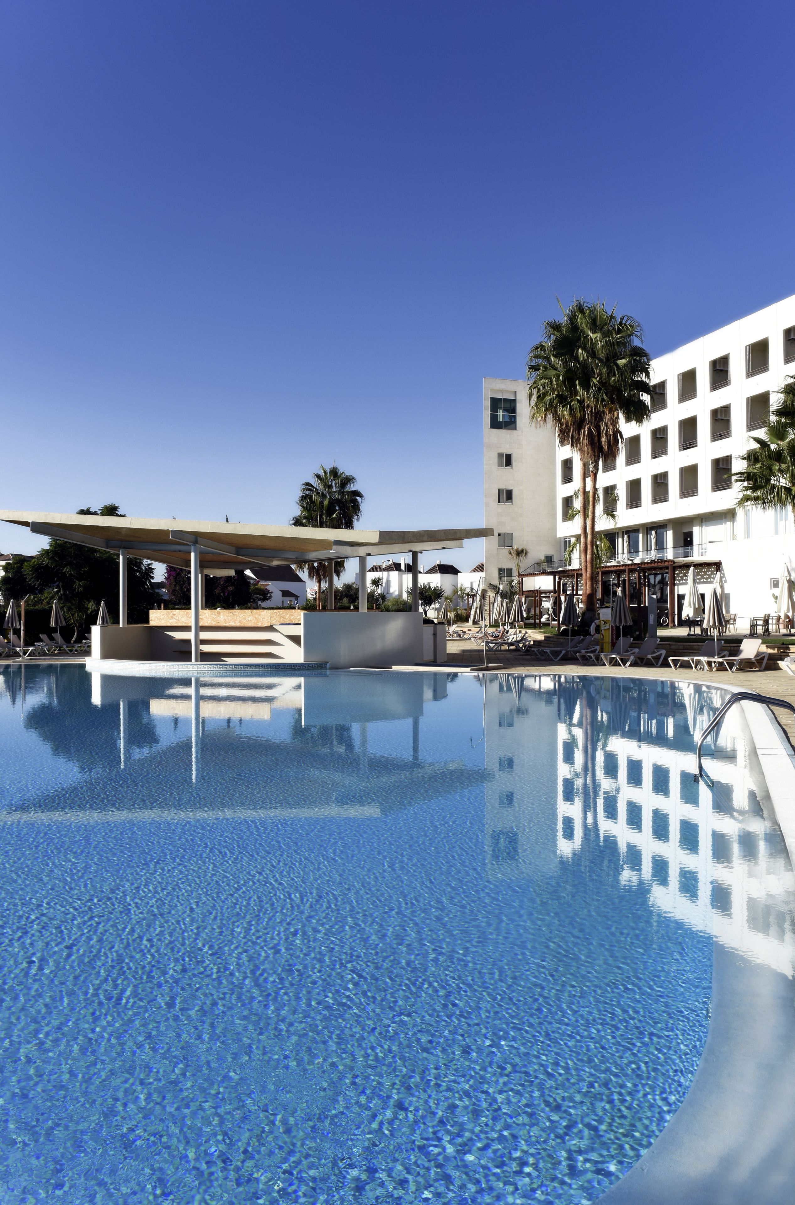 A clear blue pool with lounge chairs and umbrellas, surrounded by a white building with palm trees.