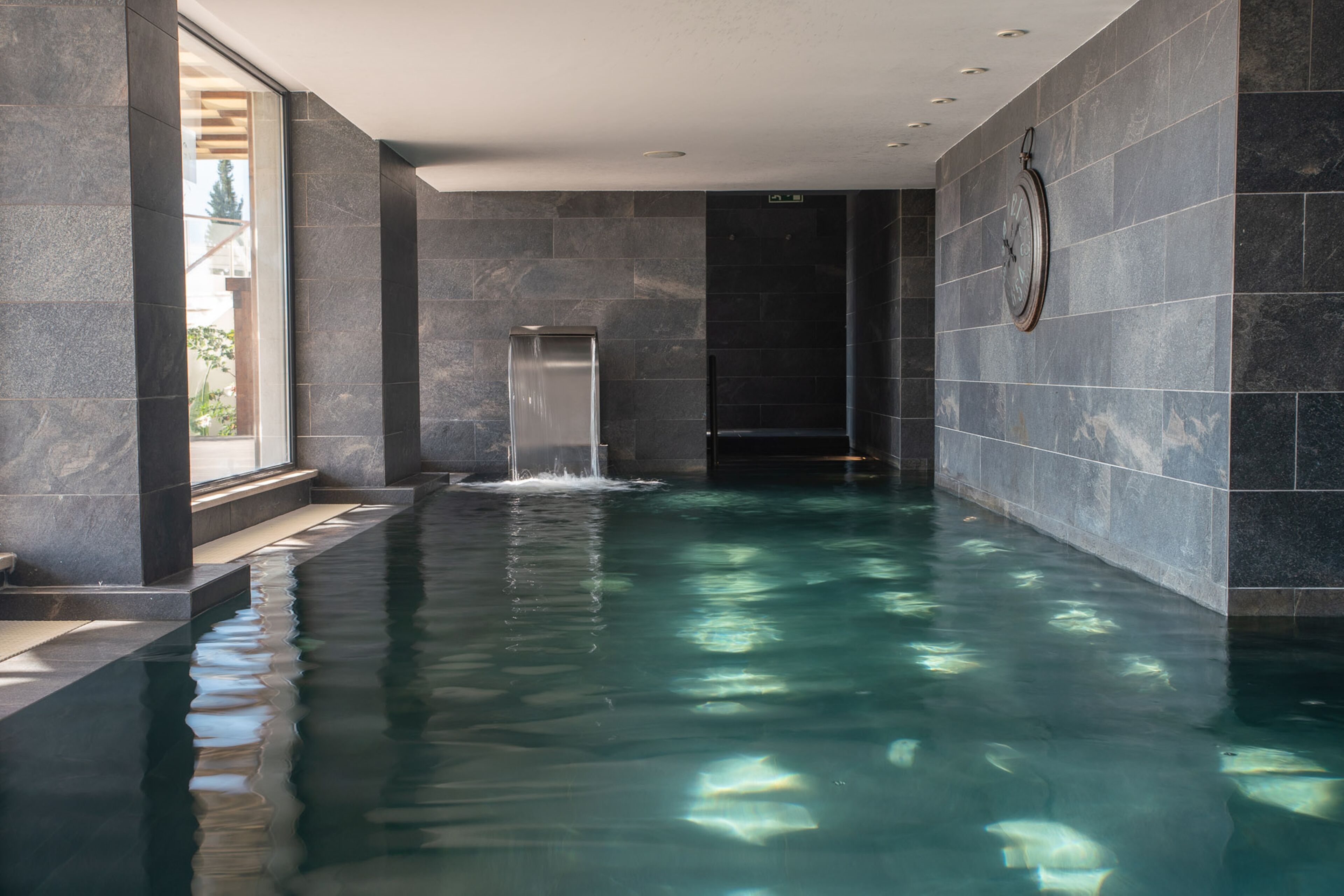 A modern indoor swimming pool with a waterfall feature, surrounded by dark stone walls and a large window.