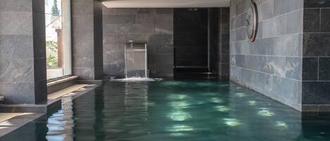 A modern indoor swimming pool with a waterfall feature, surrounded by dark stone walls and a large window.